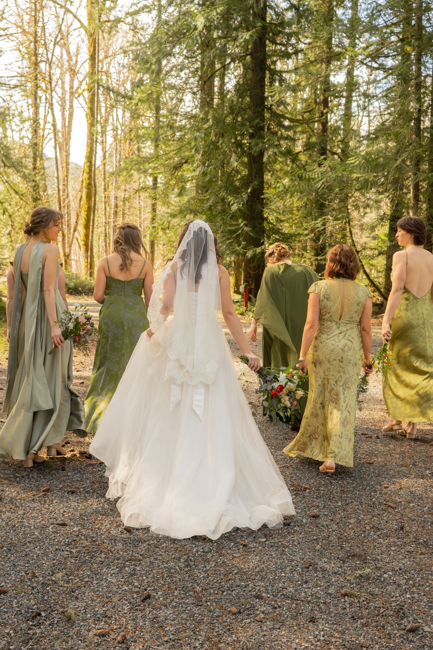 Bride in white gown with long veil walks with bridesmaids in sage green dresses through sunlit forest.