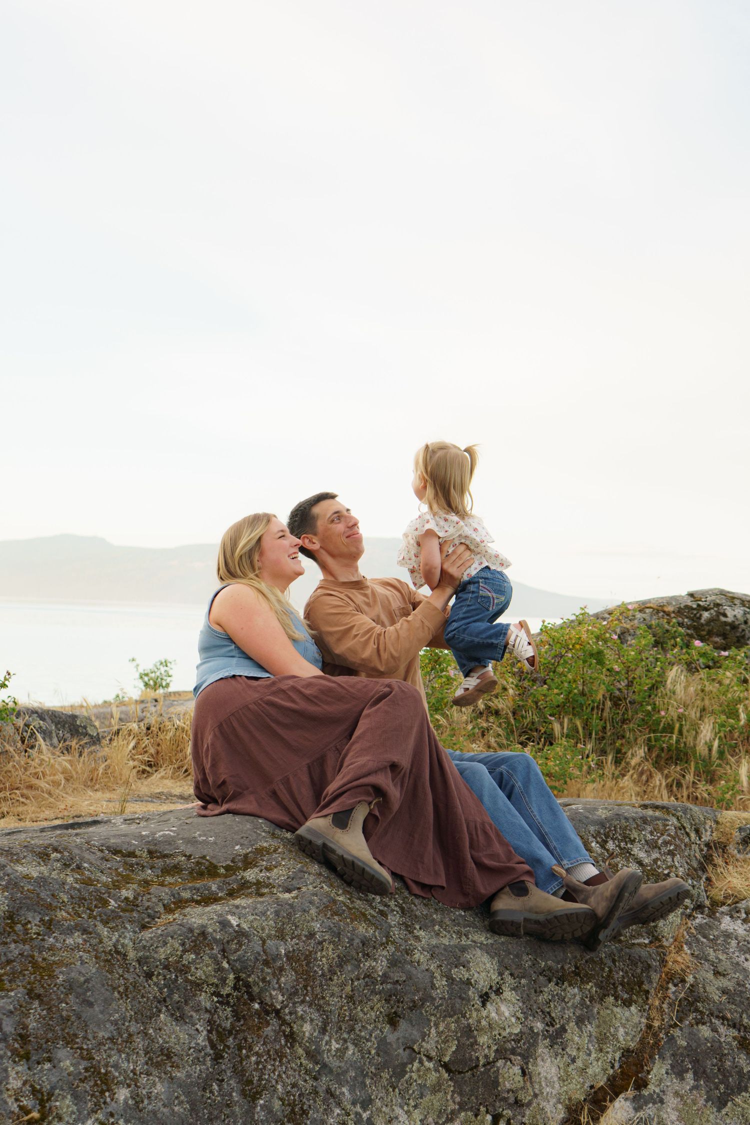 Family of three sits on a rocky hillside with scenic misty mountains in background.