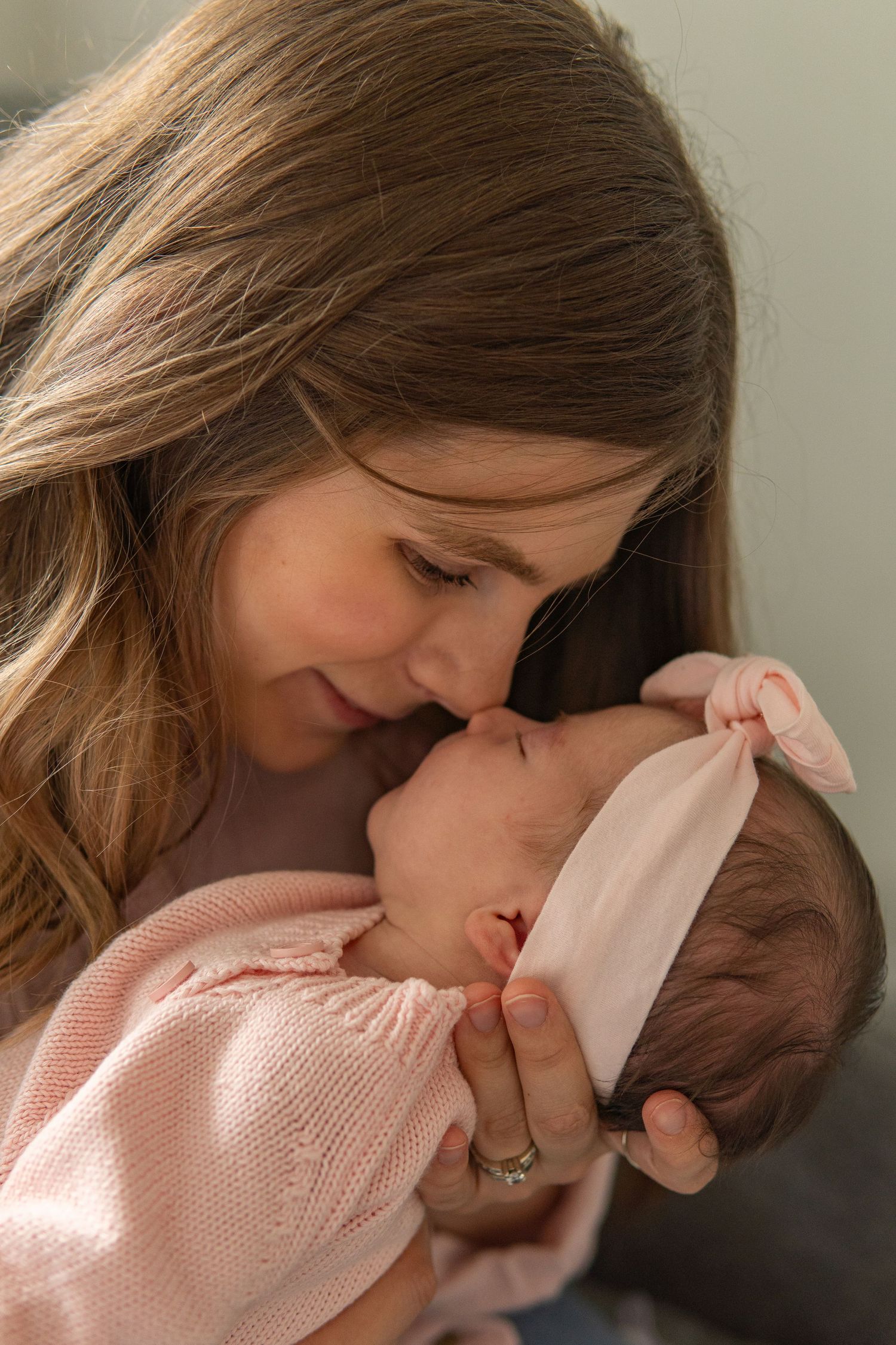 A loving mother gently touches noses with her newborn baby girl dressed in pink with a bow headband.