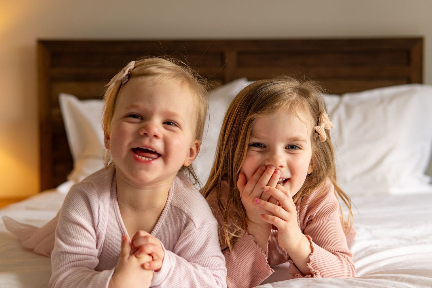 Two young girls in pink pajamas laughing and smiling together on a white bed.