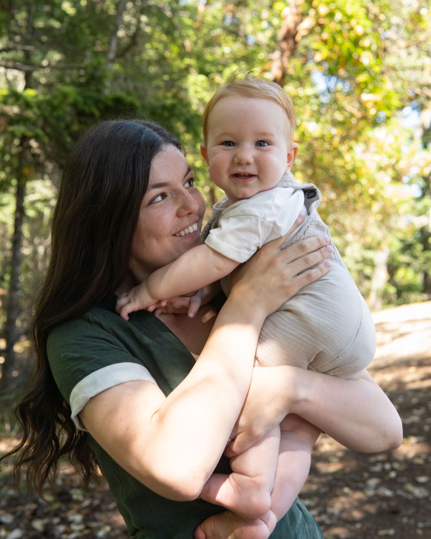 A smiling woman in a green shirt holds a happy baby in a sunlit forest setting.