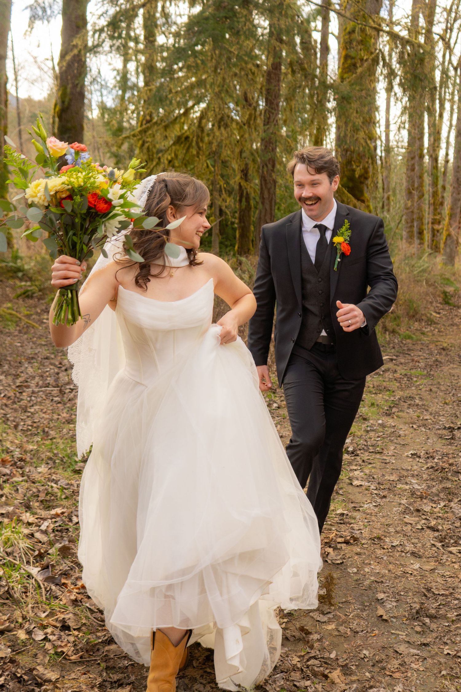 A joyful bride and groom walk hand in hand along a forest path holding a colorful bouquet.