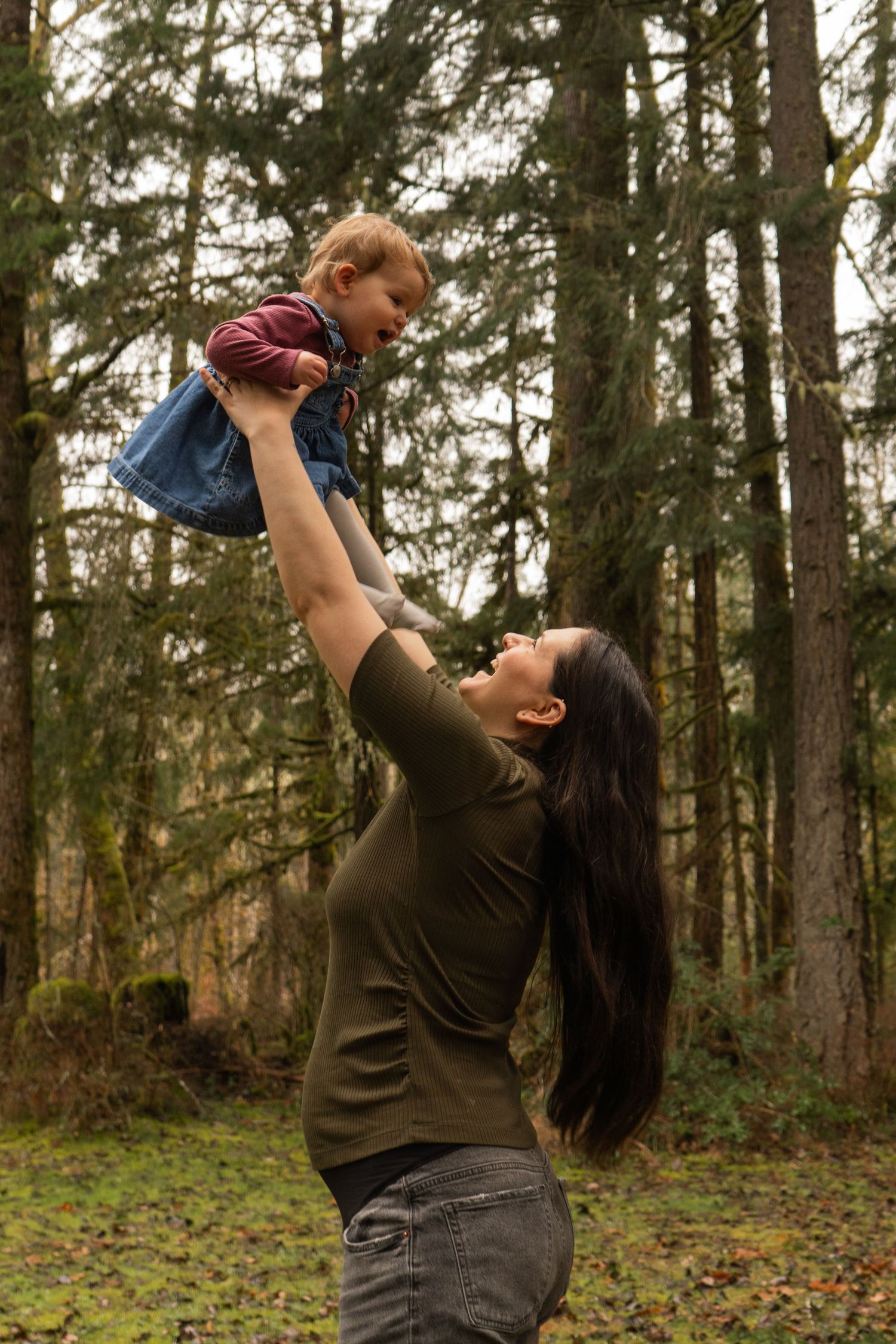 A mother joyfully lifts her toddler high in the air in a lush forest setting.