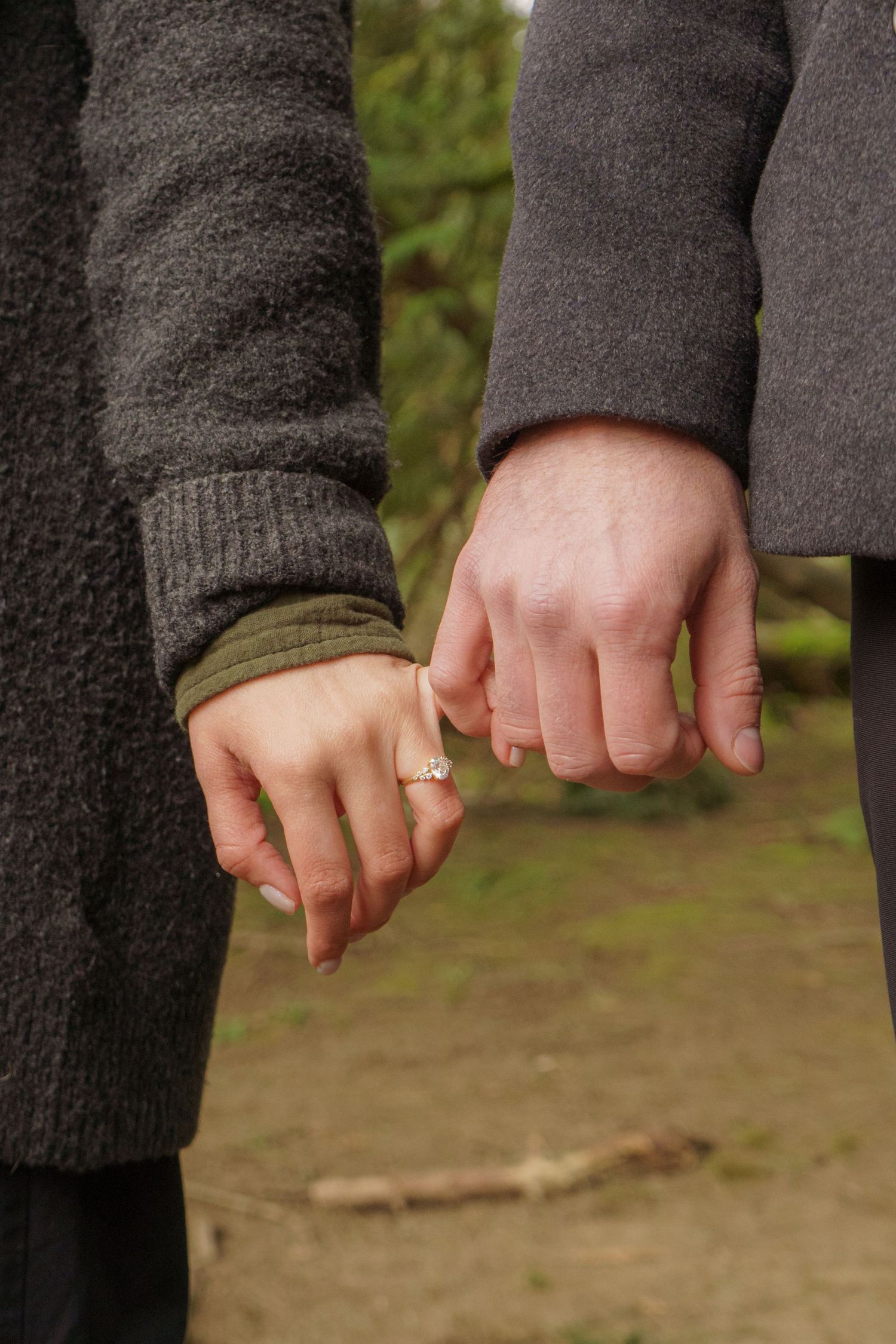Couple holding hands outdoors, woman wearing a diamond engagement ring, both dressed in gray coats.