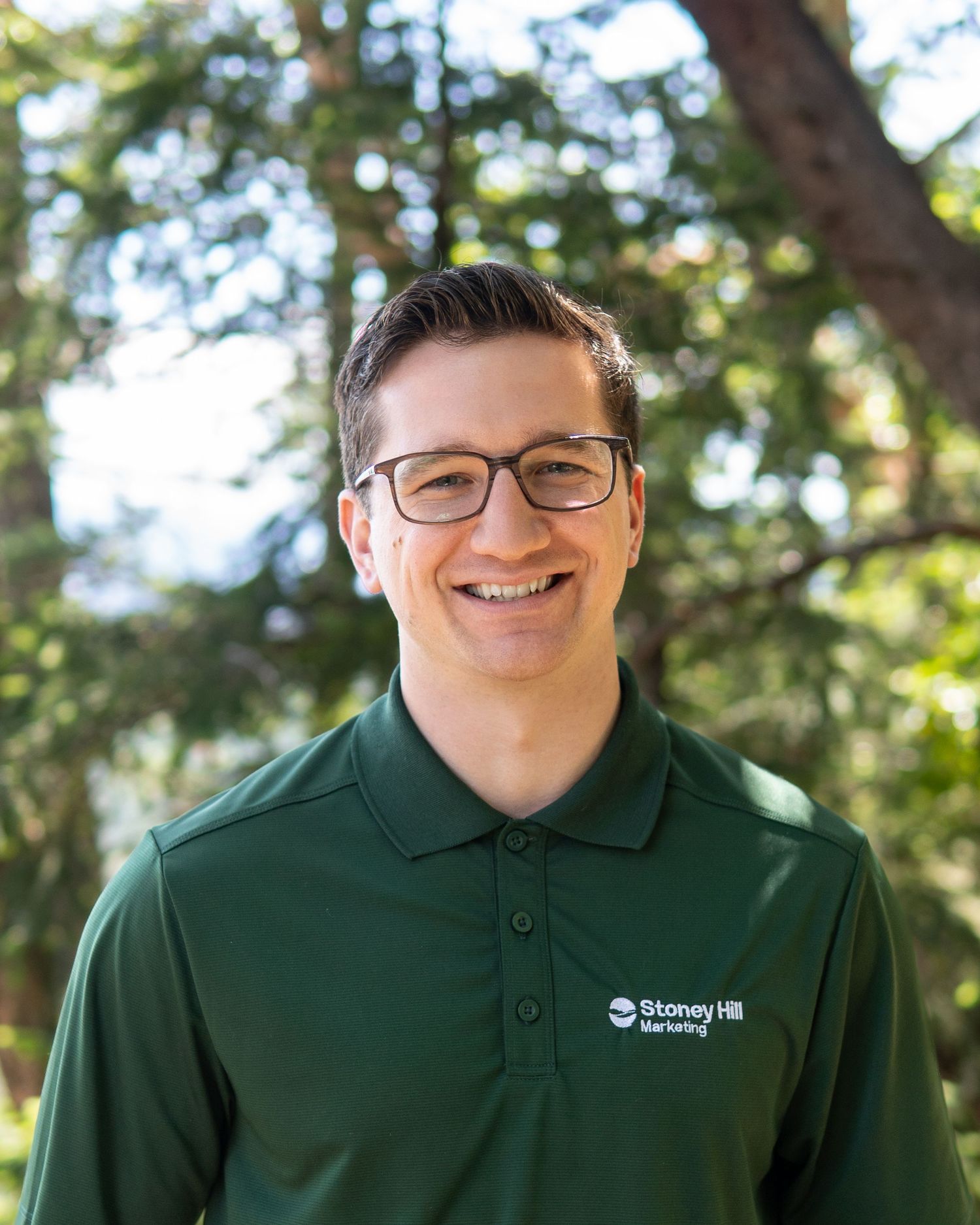 Smiling man wearing glasses and a green Stoney Hill Marketing polo shirt outdoors with trees in background.