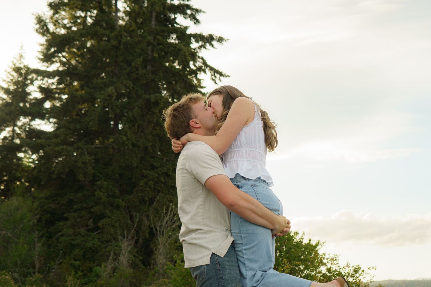 A couple shares a romantic kiss outdoors, surrounded by tall evergreen trees and a soft cloudy sky.