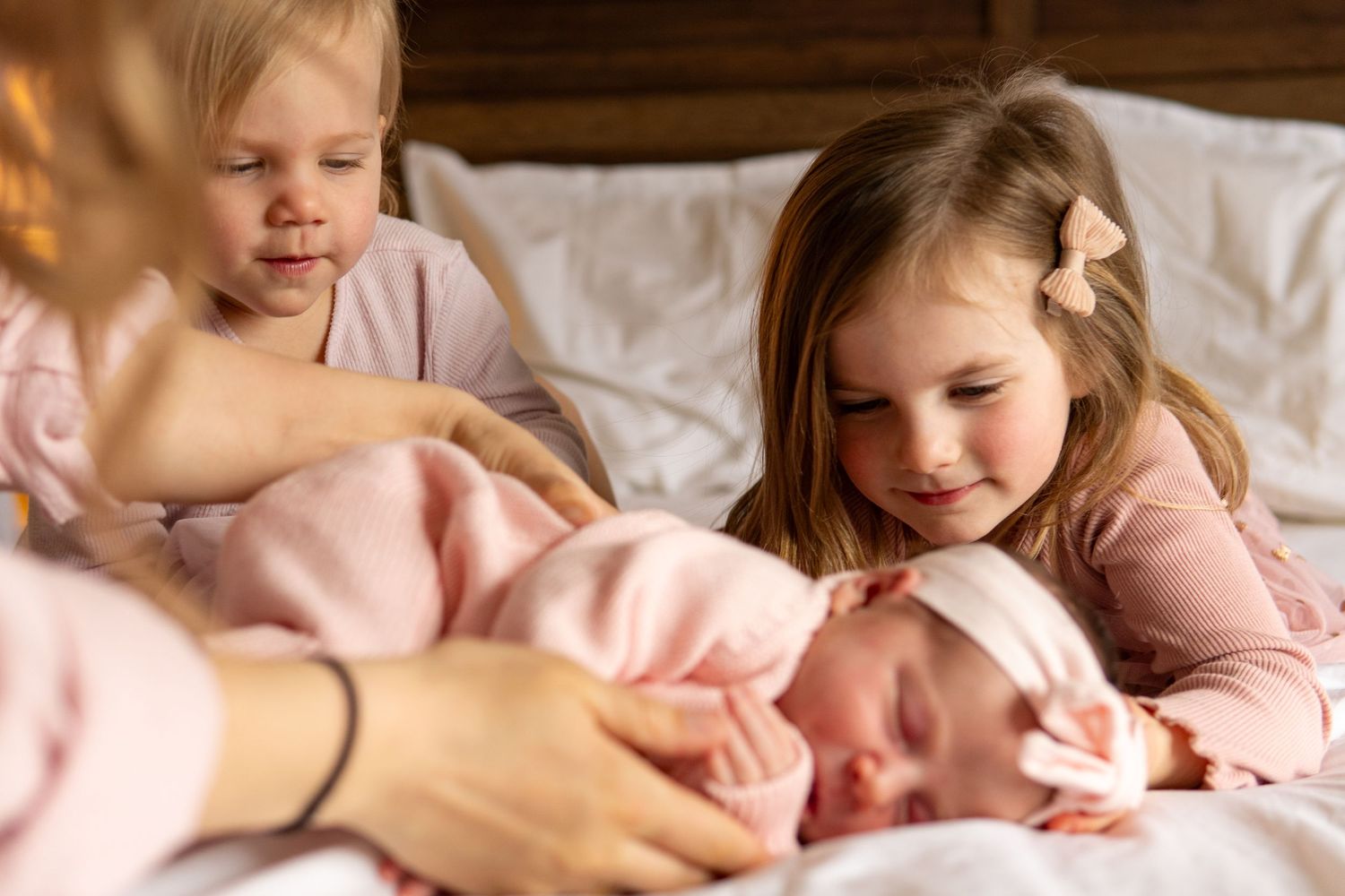 Two young siblings lovingly gaze at their newborn baby sister lying on a white bed in a cozy family moment.
