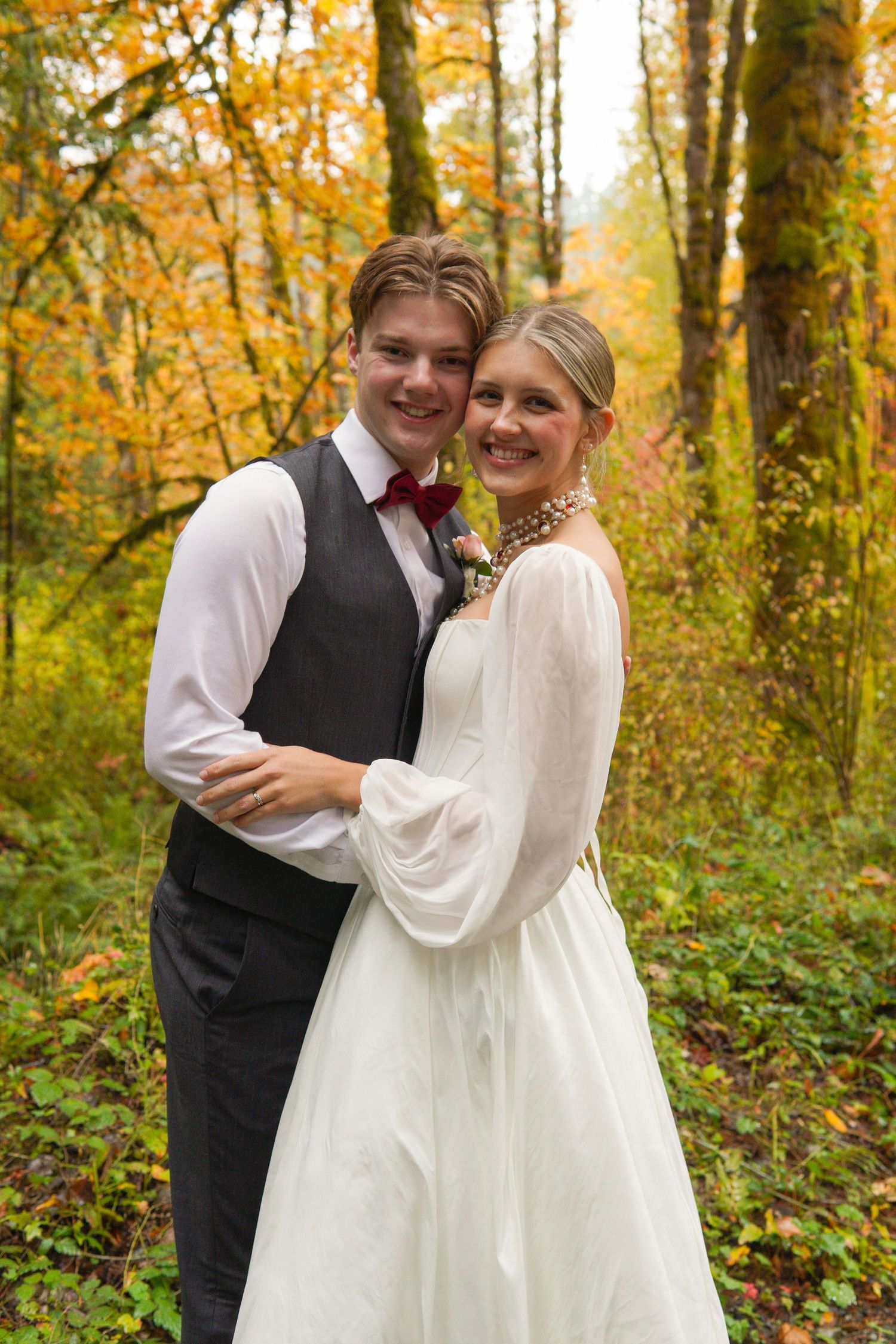 Bride and groom embracing in autumn forest, wearing white gown and dark vest with red tie, surrounded by golden fall foliage.