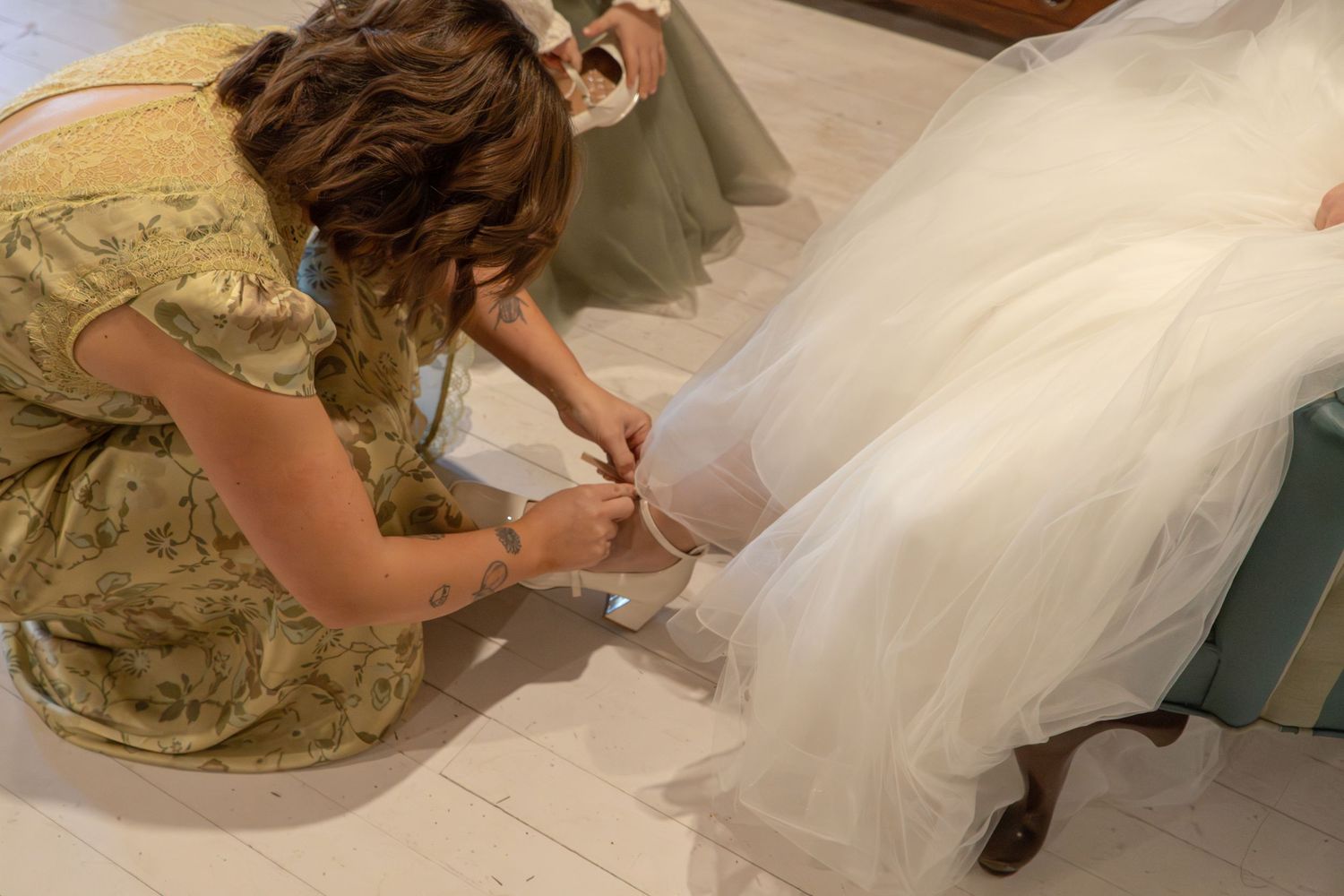 Bridesmaid in floral dress helping bride put on wedding shoes, adjusting bridal footwear before ceremony.