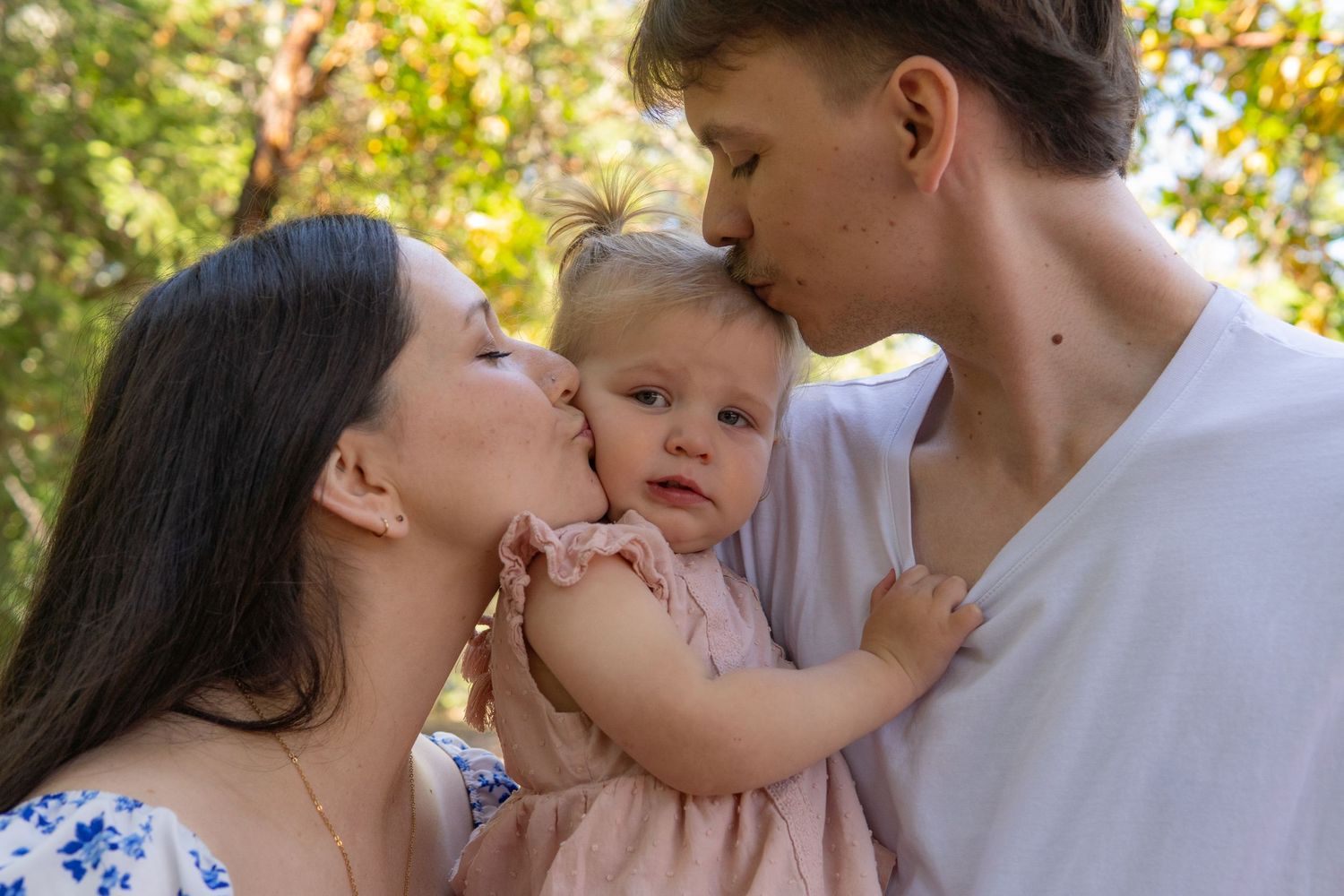 Parents kissing their baby girl on the cheeks outdoors in a sunlit park setting.