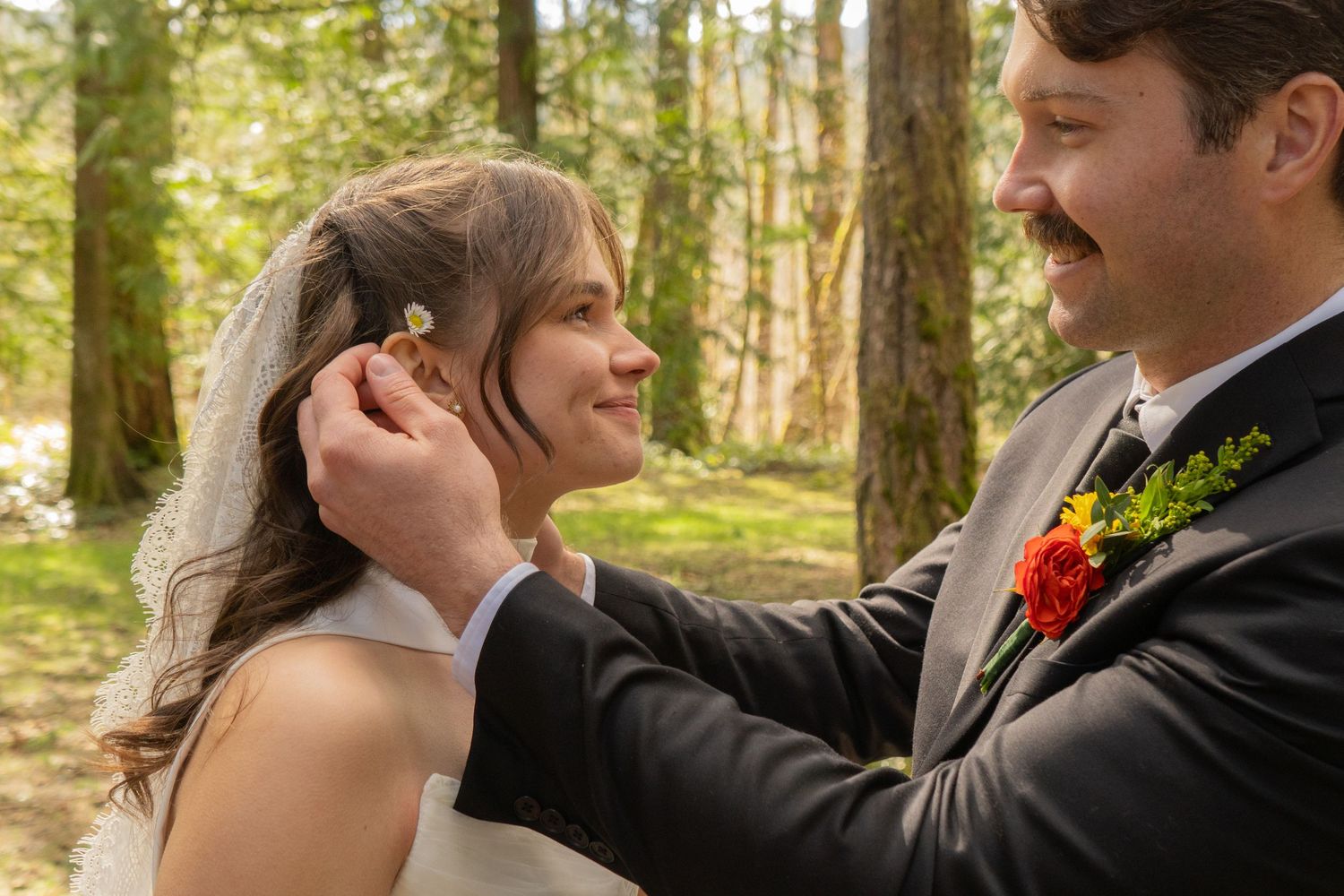 Groom tenderly places flower in bride's hair among forest trees on wedding day.