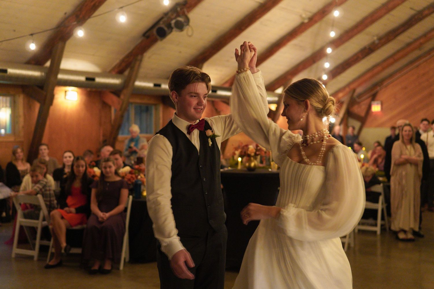 Bride and groom share first dance at rustic barn wedding reception with guests watching in background.