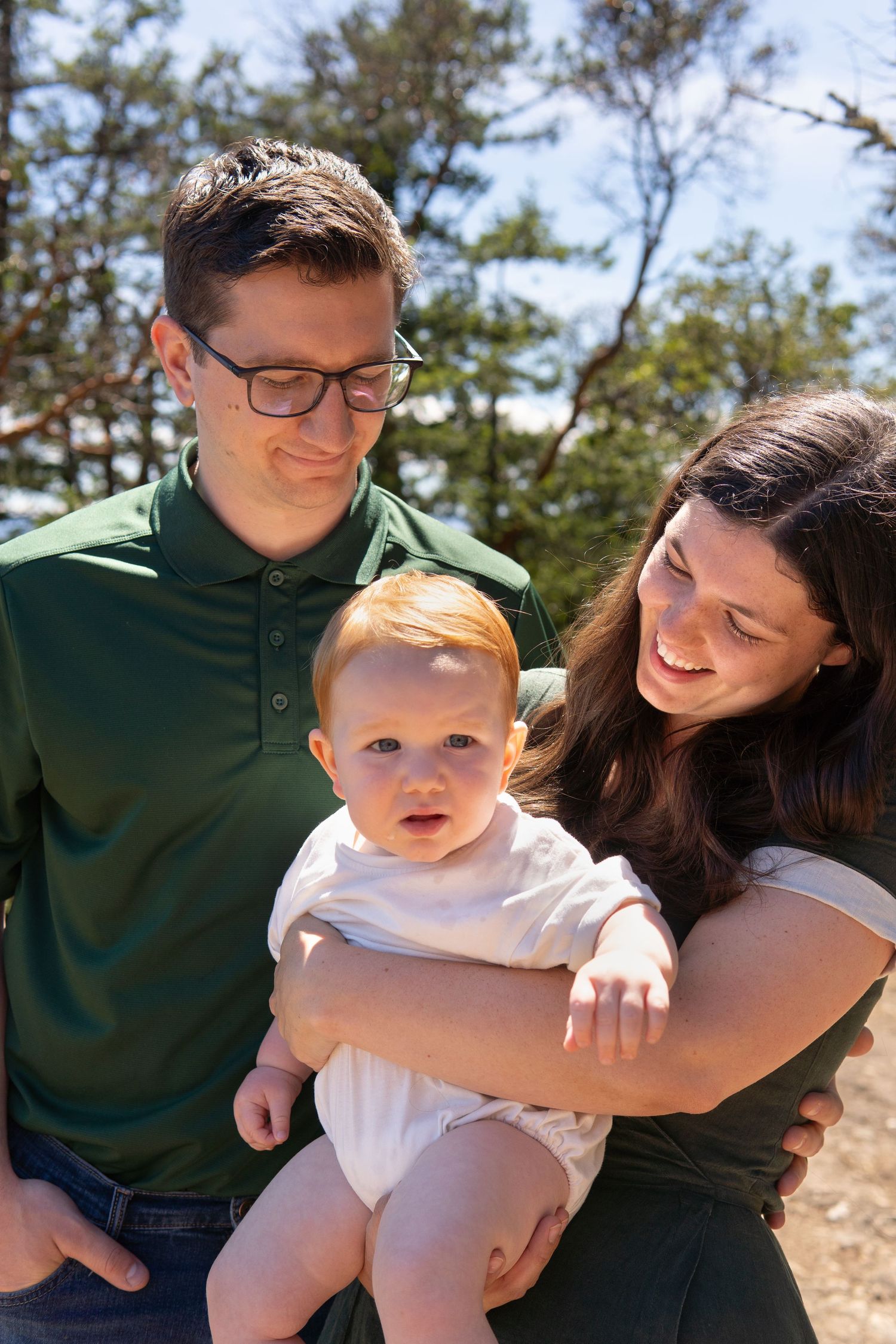 Young couple smiling outdoors holding their red-haired baby, surrounded by pine trees on a sunny day.