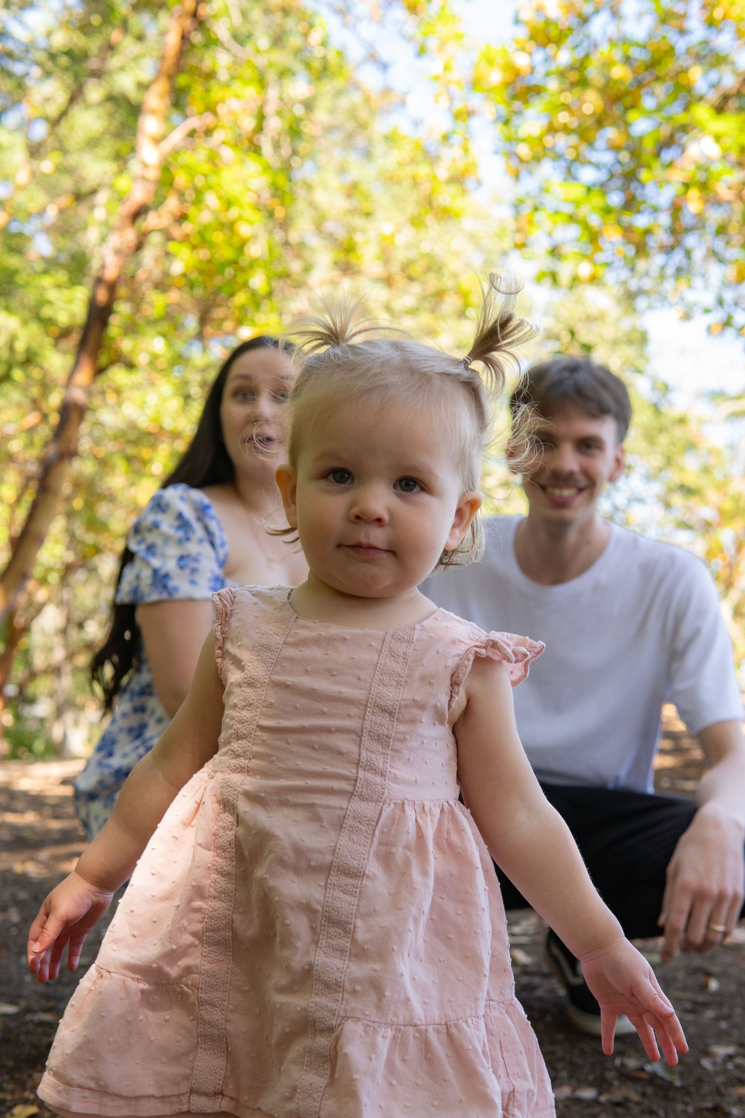 Toddler girl in pink dress stands in forest with smiling parents seated behind her on sunny day.