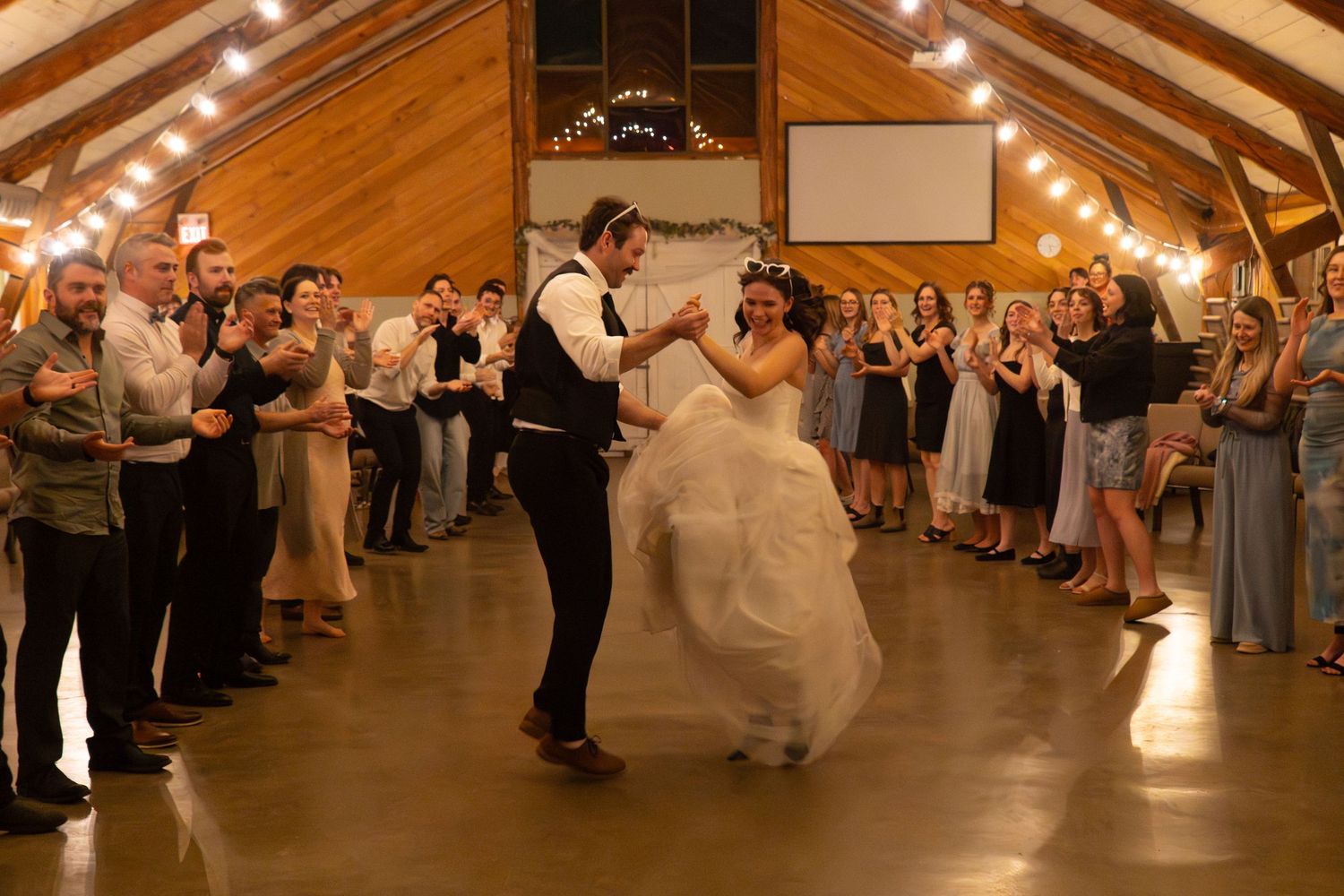 Bride and groom share their first dance in a rustic wooden barn venue surrounded by applauding wedding guests.