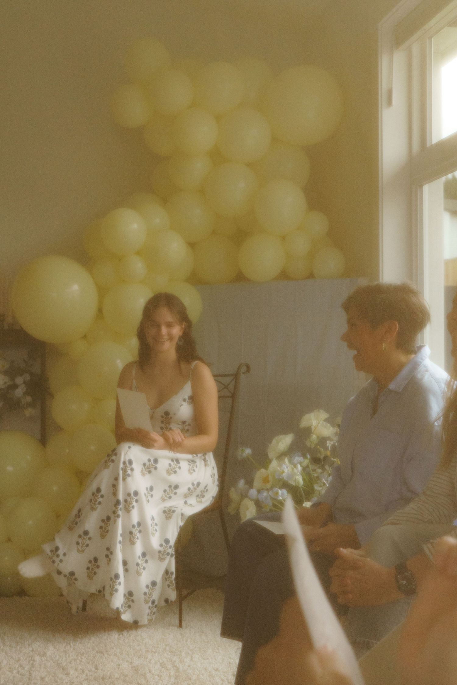 A smiling woman in a floral dress sits with a man holding flowers amid yellow balloons.