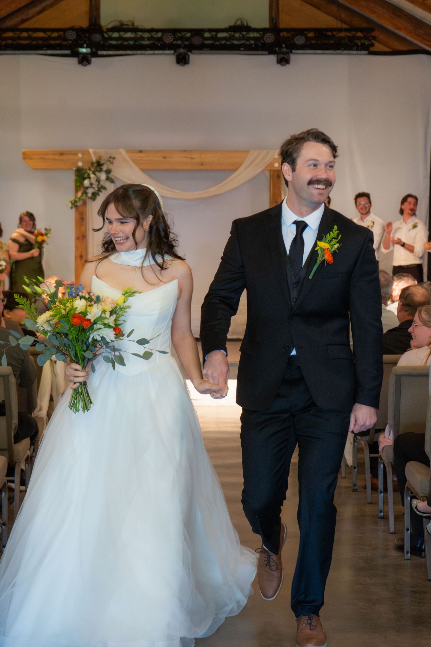Newlywed couple walks down the aisle hand-in-hand, bride in white gown holding colorful bouquet, groom in black suit.