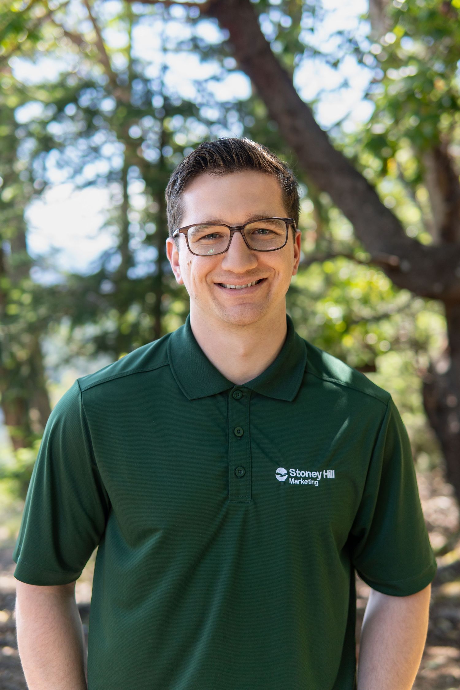 Smiling man with glasses wearing a green Stoney Hill Marketing polo shirt outdoors with trees in background.