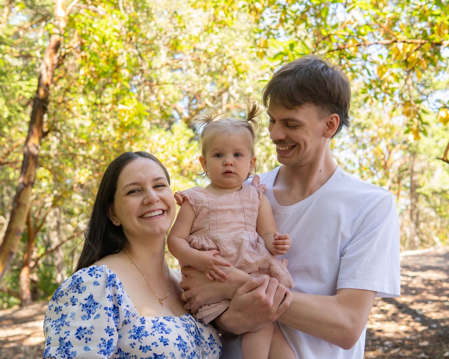 Happy young family with baby girl posing outdoors in a sunny green forest setting.