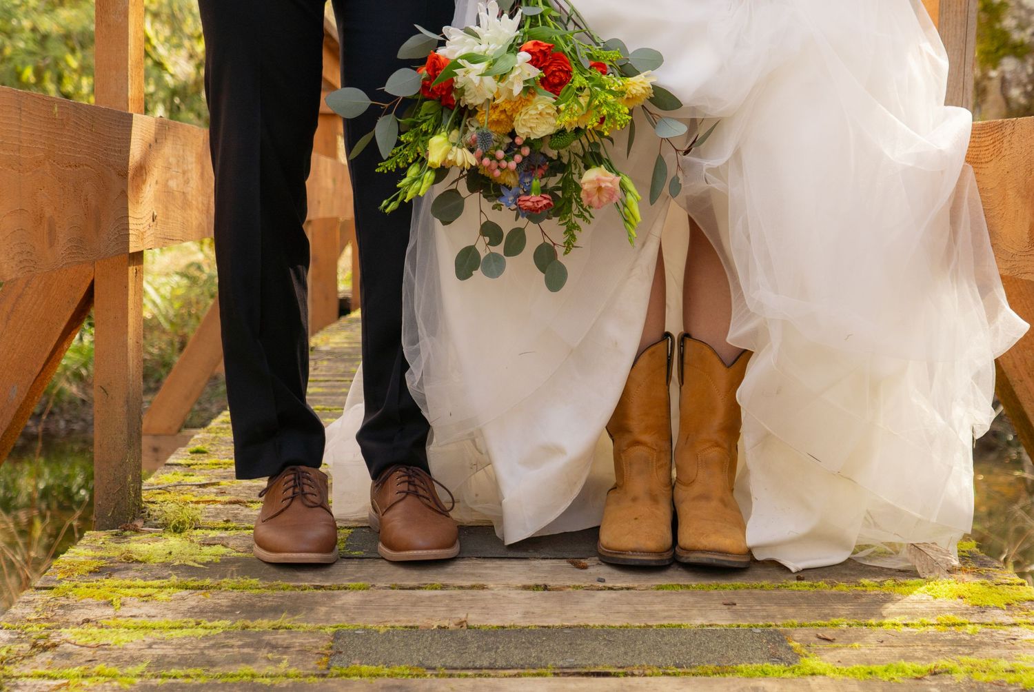 Bride in cowboy boots and groom in brown dress shoes standing on wooden bridge holding colorful wedding bouquet.