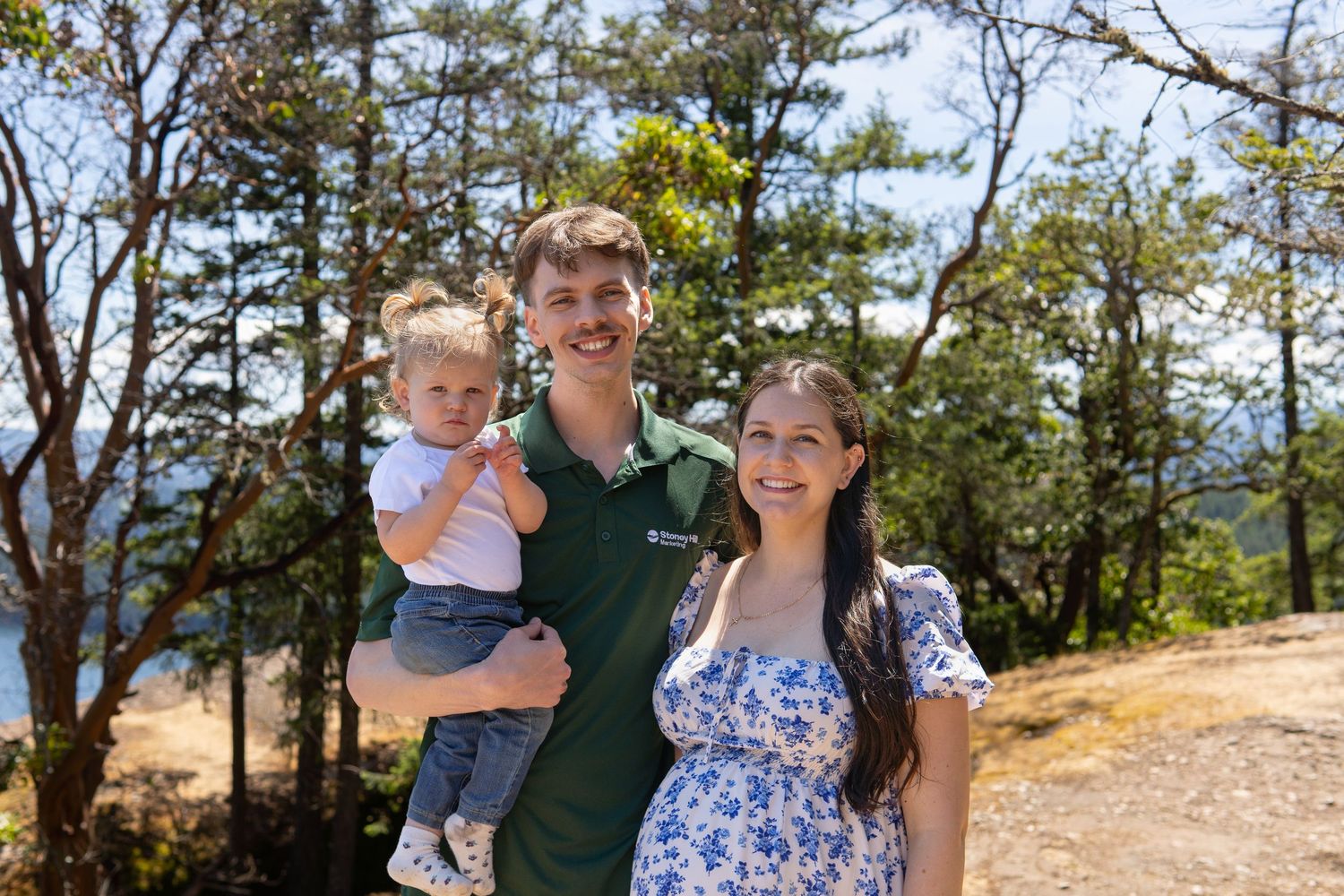 A young family smiling outdoors among pine trees, with a man holding a baby and a pregnant woman beside him.