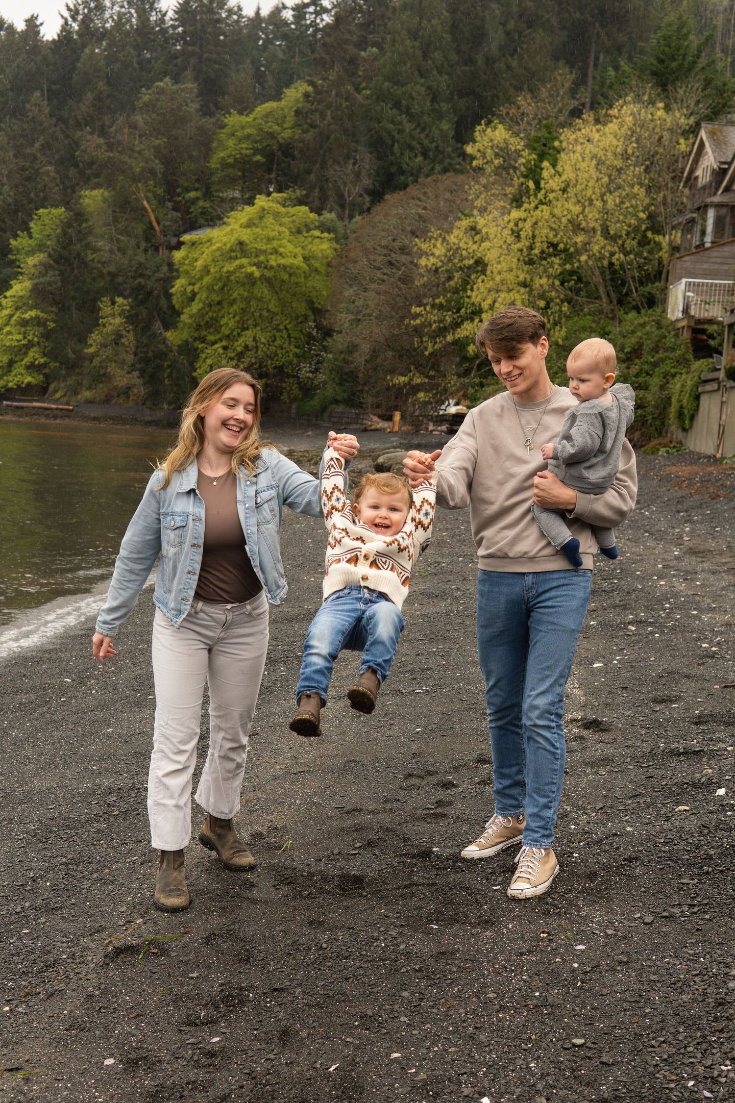 A young family laughs while swinging a toddler on a rocky beach, with lush green trees and a calm bay behind them.