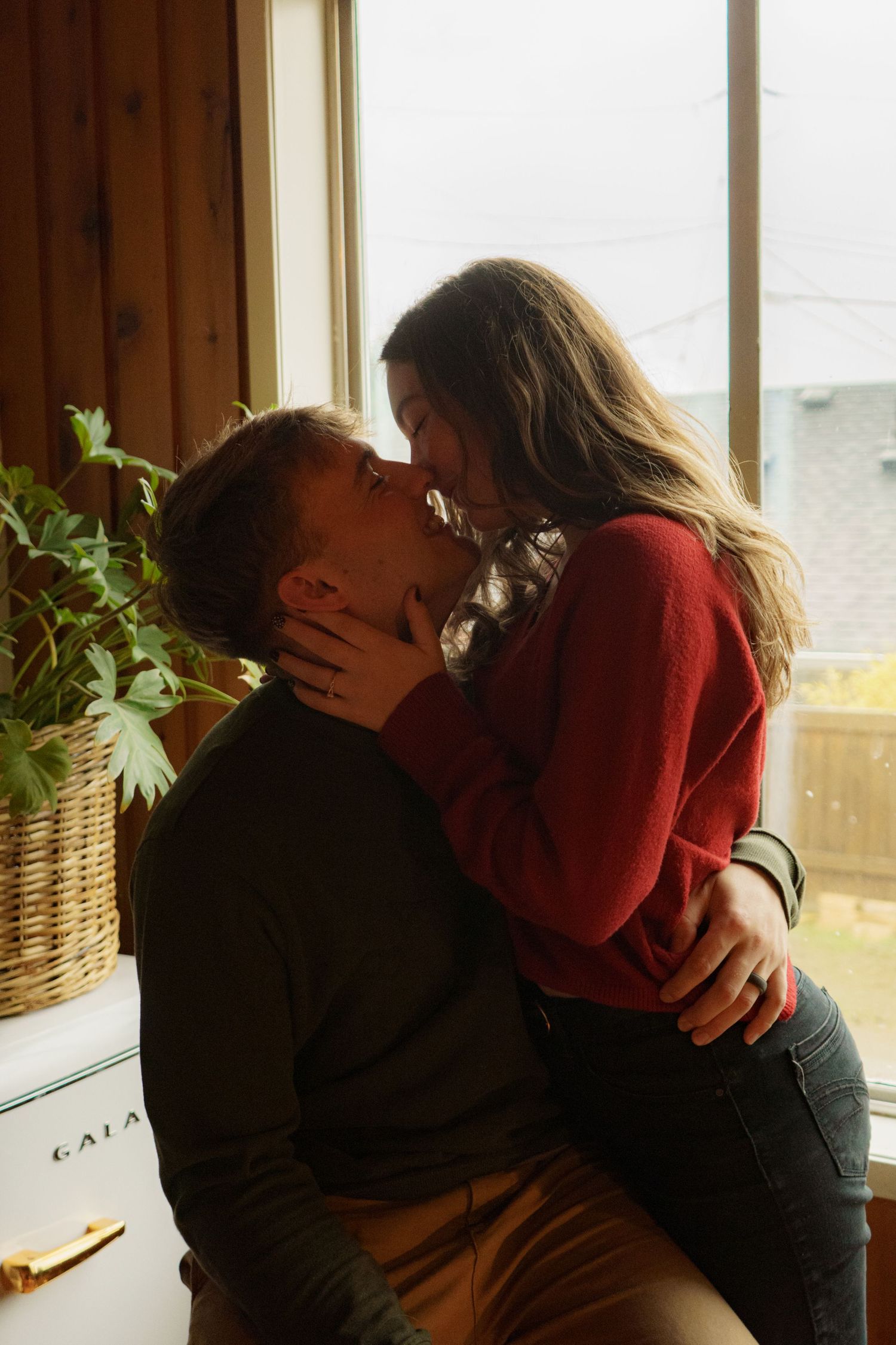 Couple sharing a passionate kiss while sitting by a sunlit window with houseplants nearby.