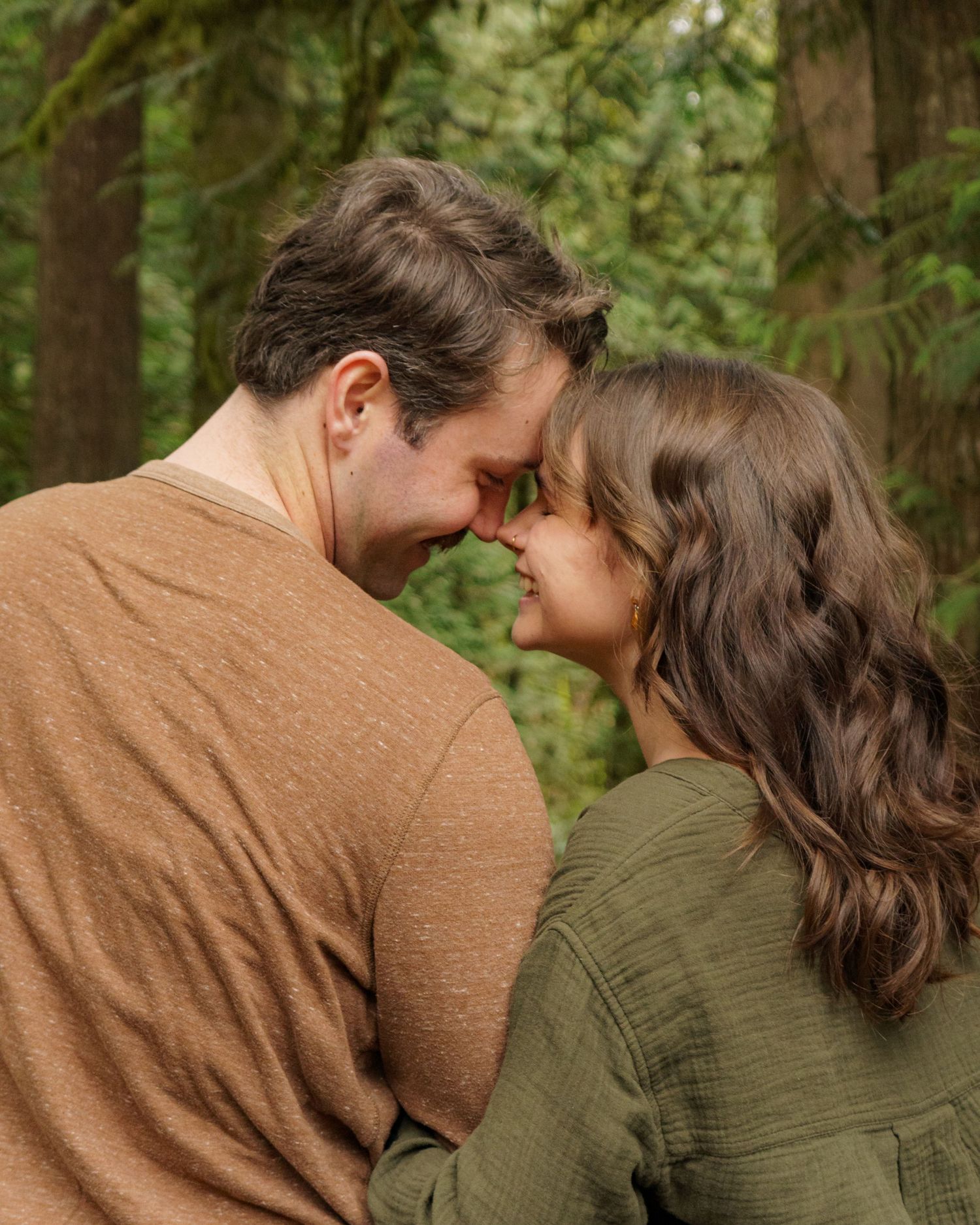 A couple shares a romantic forehead touch surrounded by lush green forest trees in a tender outdoor moment.