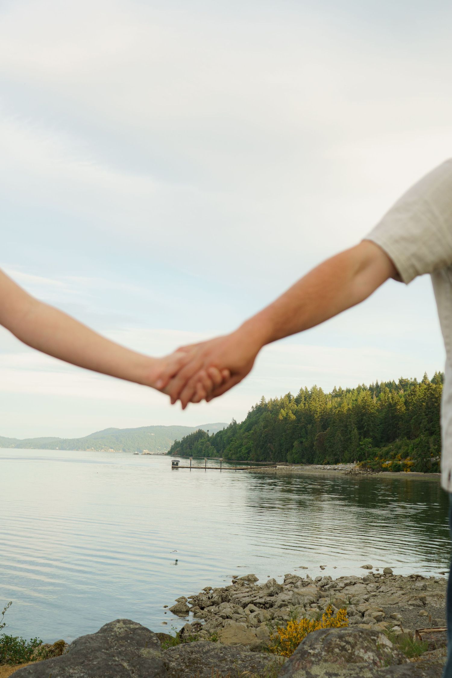Couple holding hands by a serene lake with lush green forested hills and rocky shoreline under a cloudy sky.
