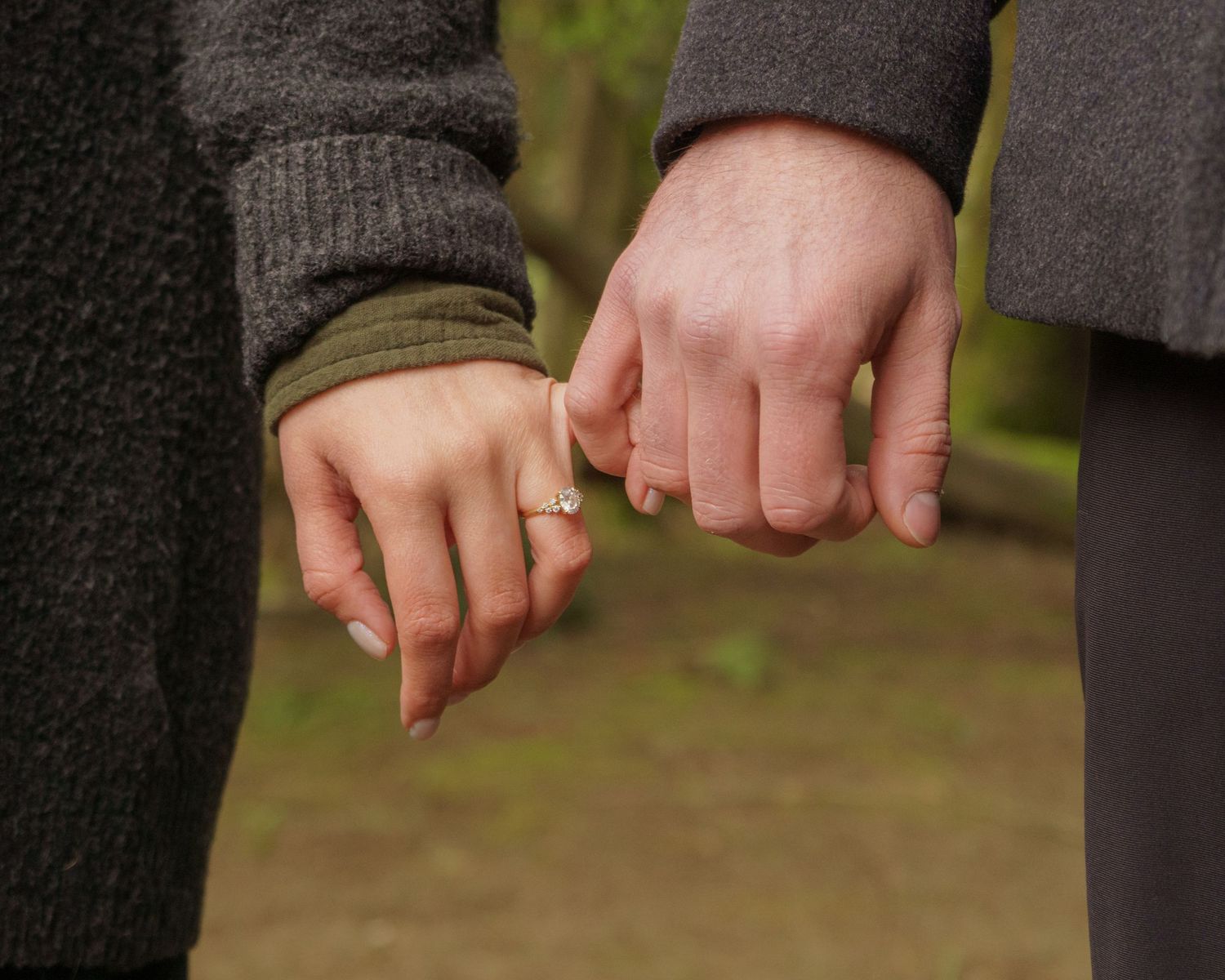 Couple holding pinkies with engagement ring visible, romantic outdoor photo.