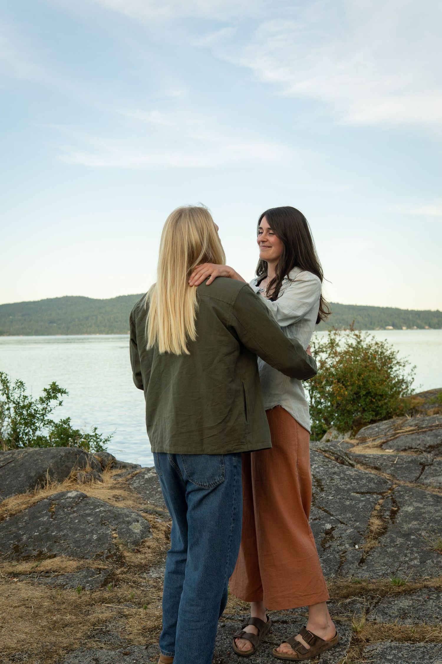 Two women embracing on rocky shoreline with scenic lake and hills in background on a cloudy day.