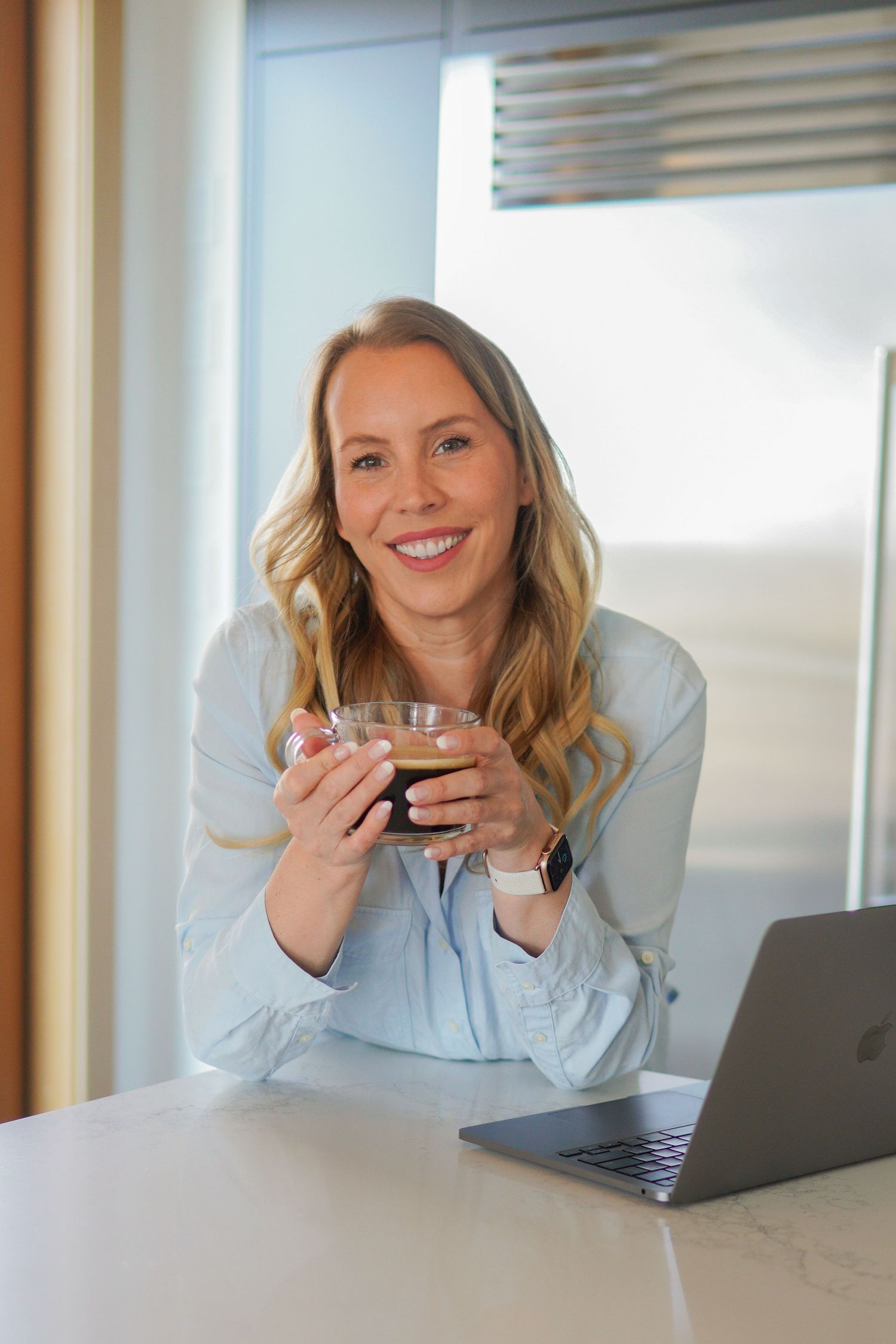 Smiling blonde woman holding a glass cup of tea at a white kitchen counter with an open laptop nearby.