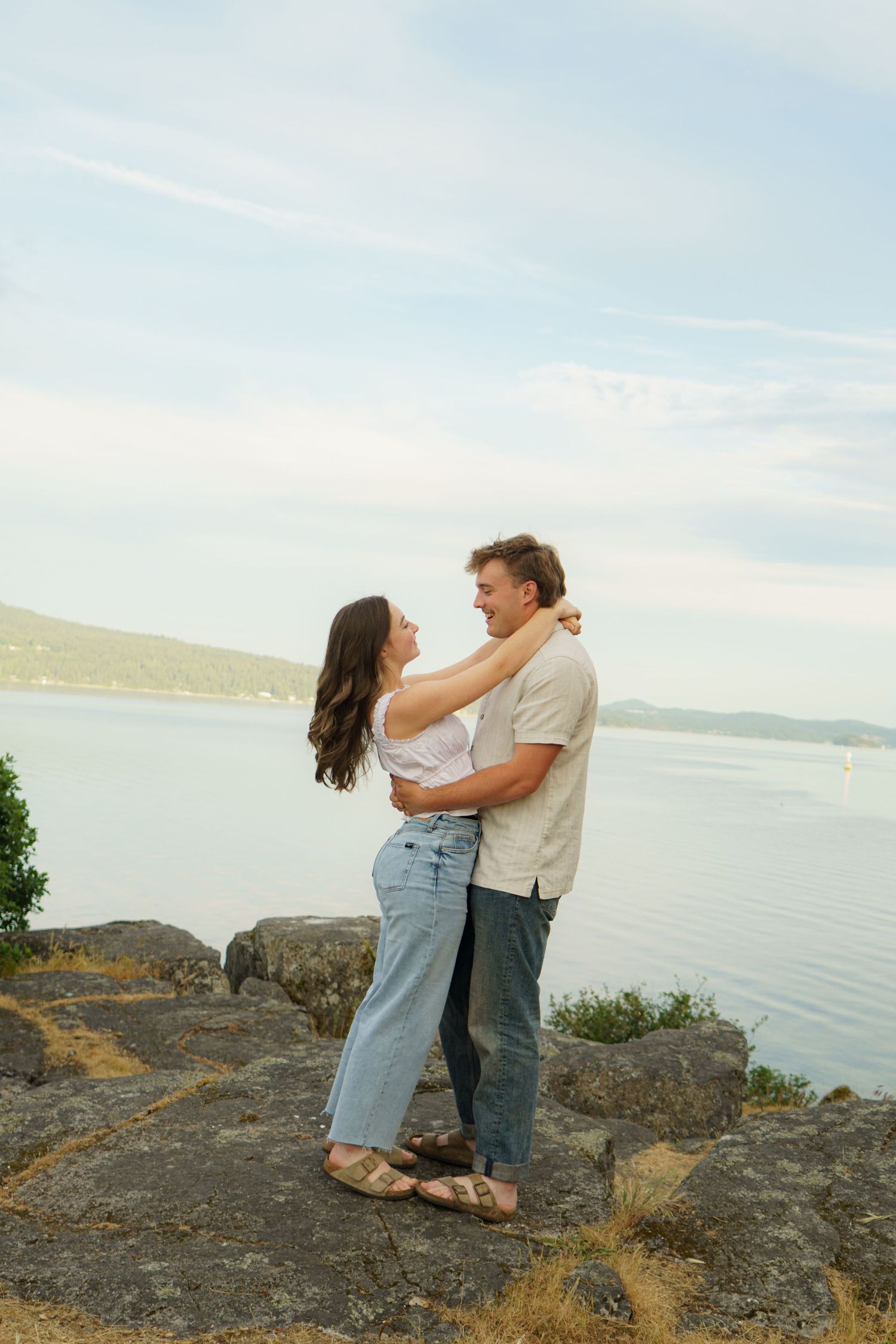 A couple embraces on rocky shoreline with scenic water and hills backdrop on a cloudy day.