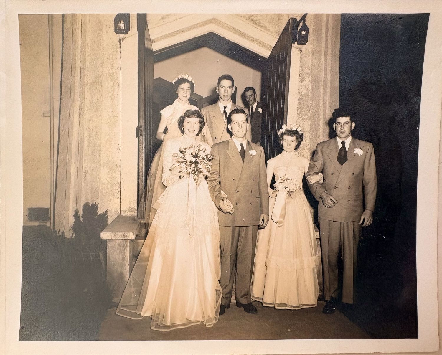 Vintage 1940s wedding party posed at church entrance, bride in white gown with bouquet, groomsmen and bridesmaids flanking couple.