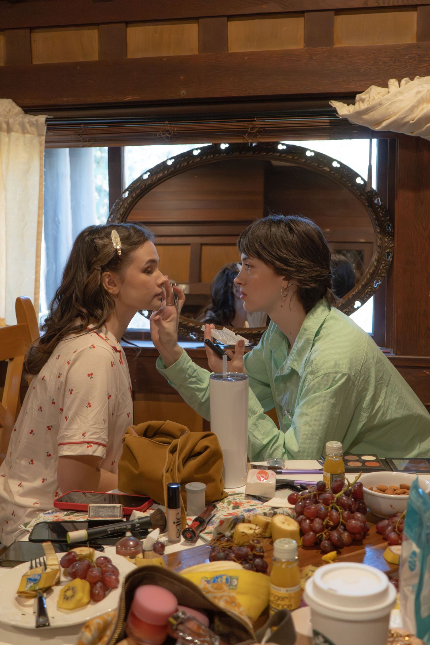 Makeup artist applying lipstick to a model at a vanity table with grapes and cosmetics, reflected in a round mirror.