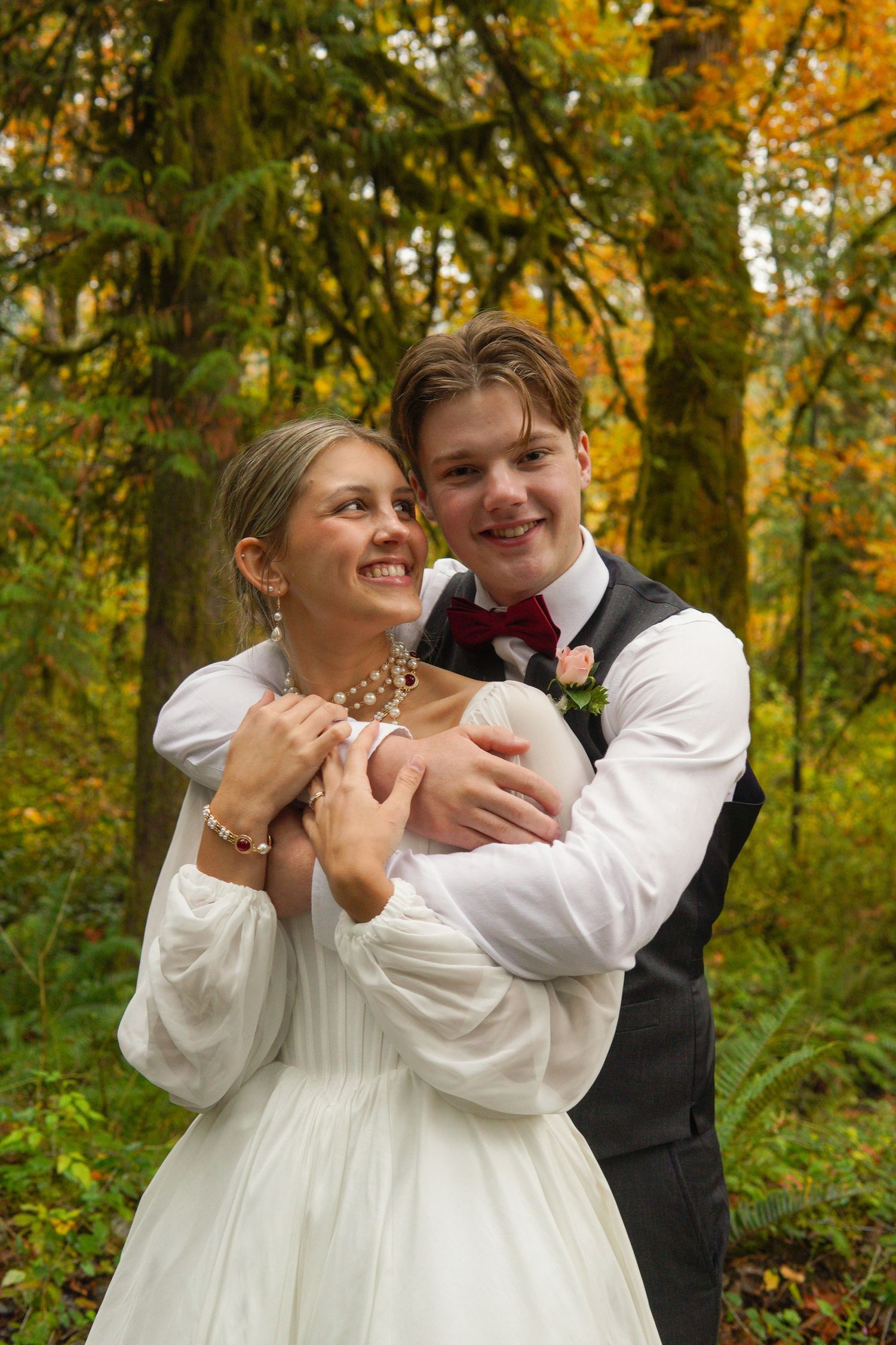 Happy newlyweds embrace in autumn forest, bride in white gown and groom in vest with pink boutonniere.