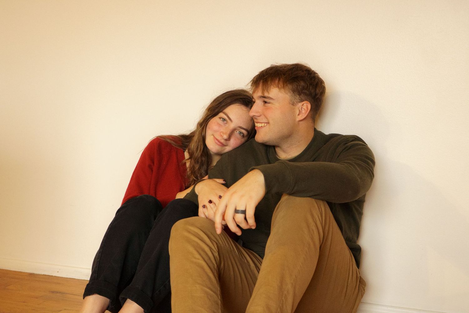 Young couple sitting on a hardwood floor leaning against a white wall holding hands.