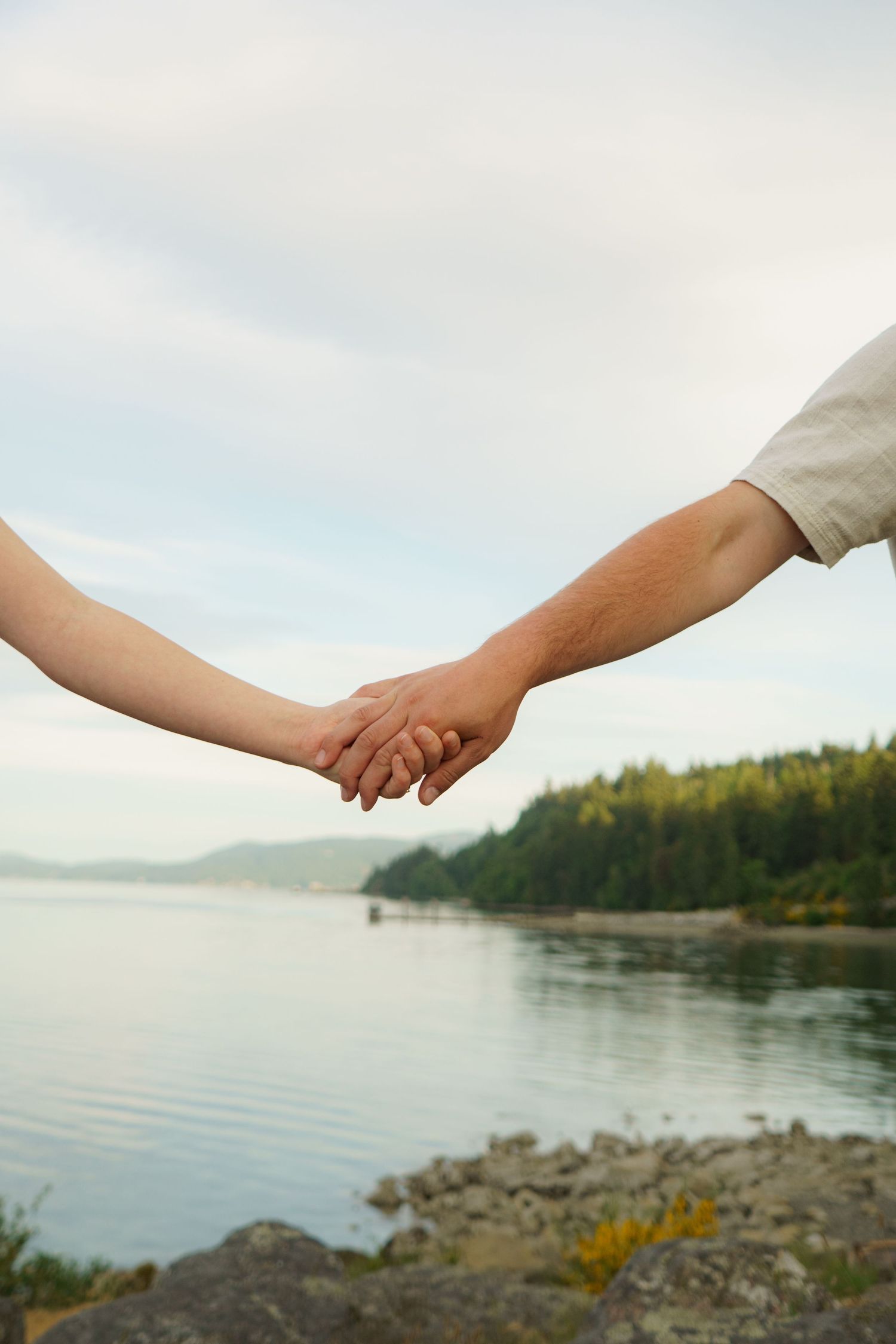 Couple holding hands by a serene lake with lush green forest and rocky shoreline in the background.