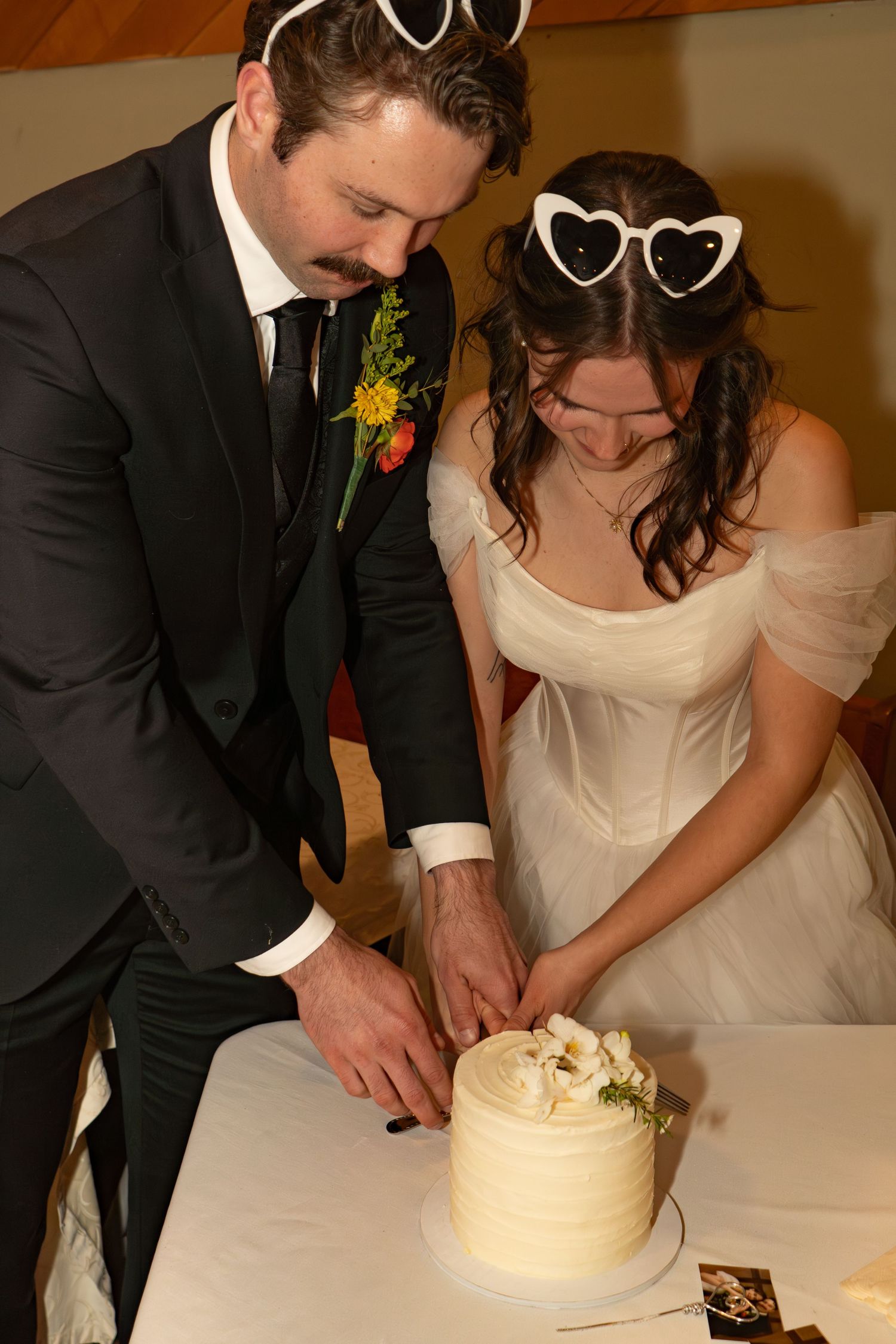 Bride and groom cutting white wedding cake together, bride wearing heart-shaped sunglasses, groom in black tuxedo.