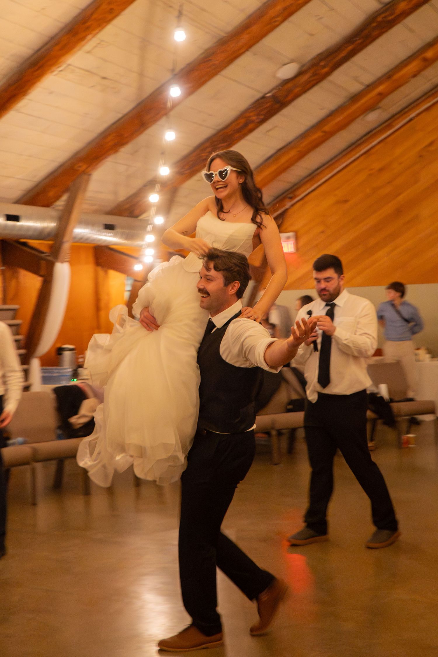 Groom lifts smiling bride in sunglasses during lively indoor wedding reception dance.
