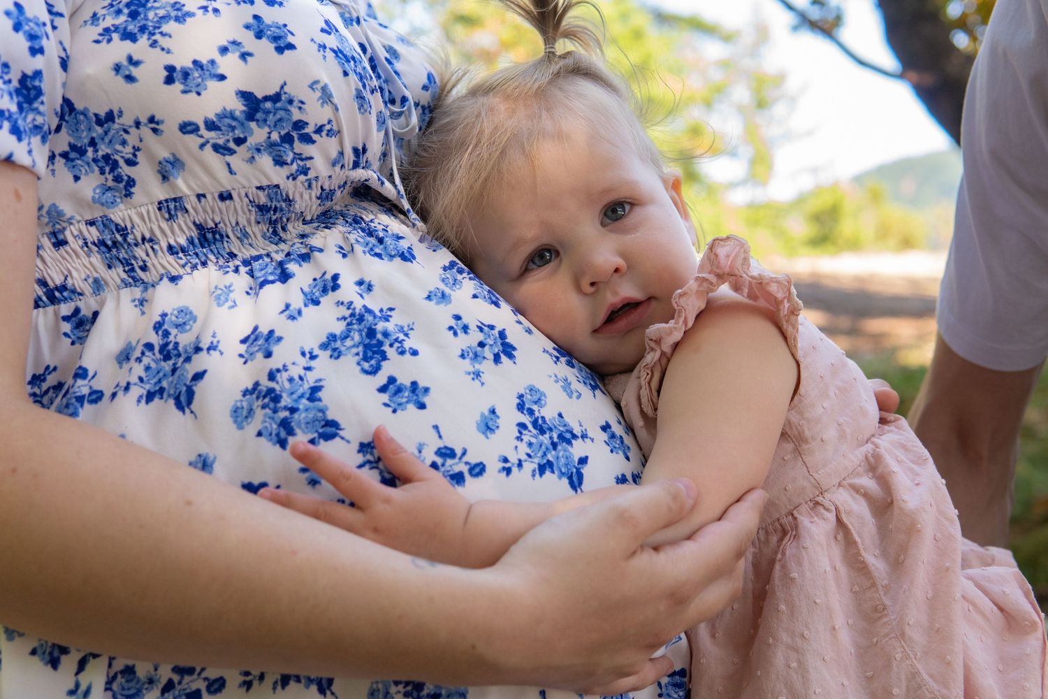 A baby girl in a pink dress rests against a pregnant woman in a blue floral dress outdoors.