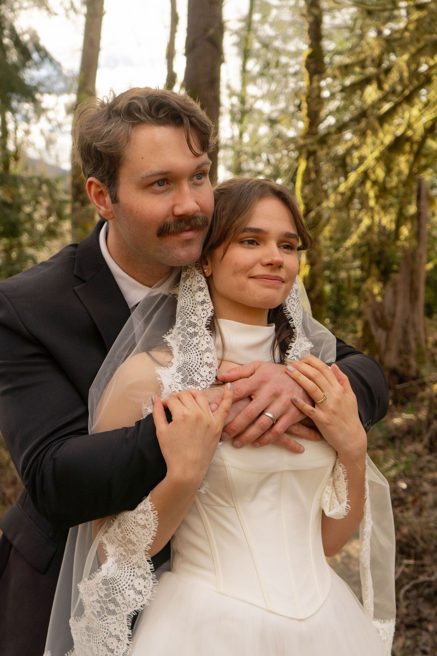 Groom embraces bride from behind in forest wearing black suit with lace veil.
