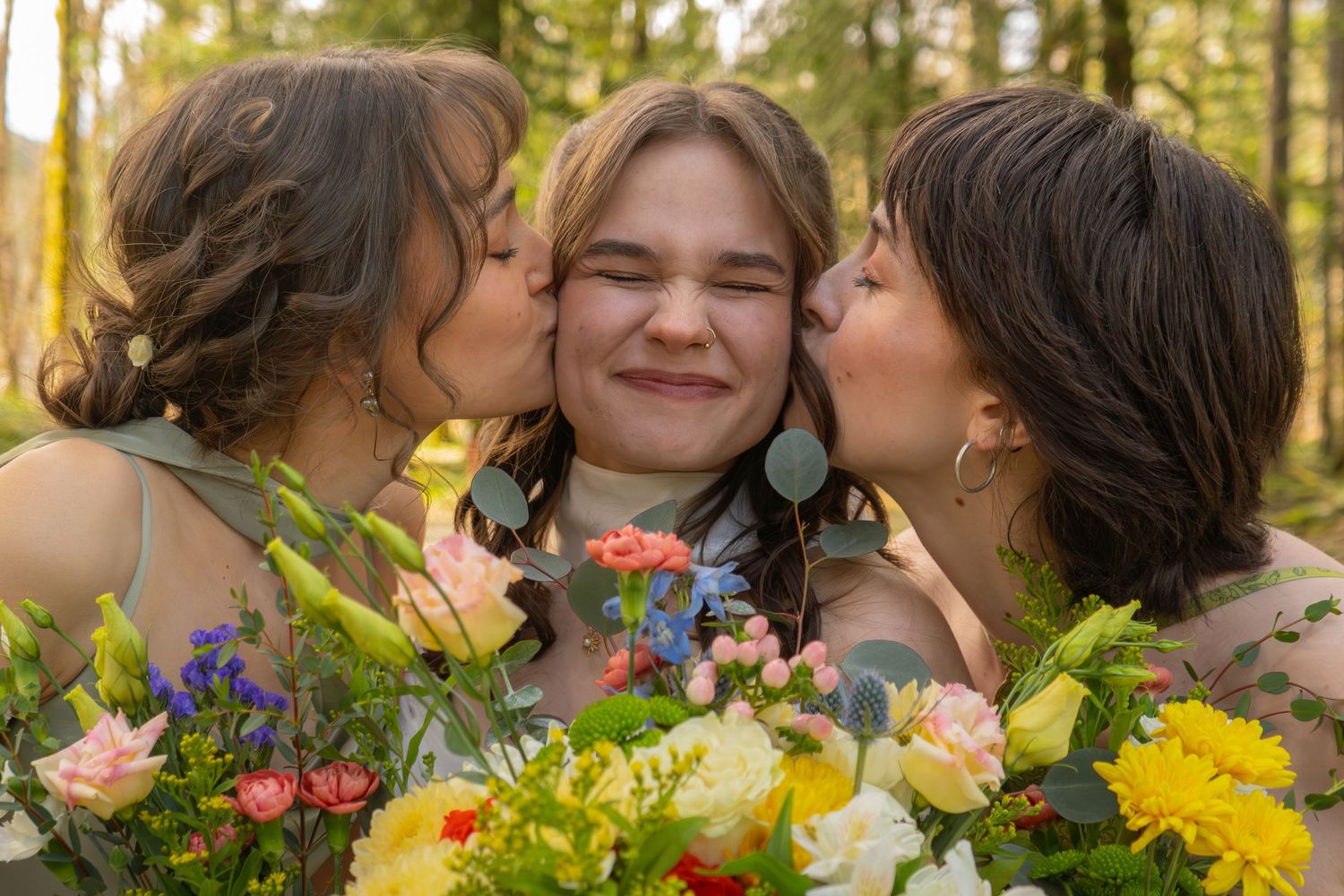 Three women celebrate outdoors with colorful bouquets, two kissing the smiling middle woman on her cheeks.