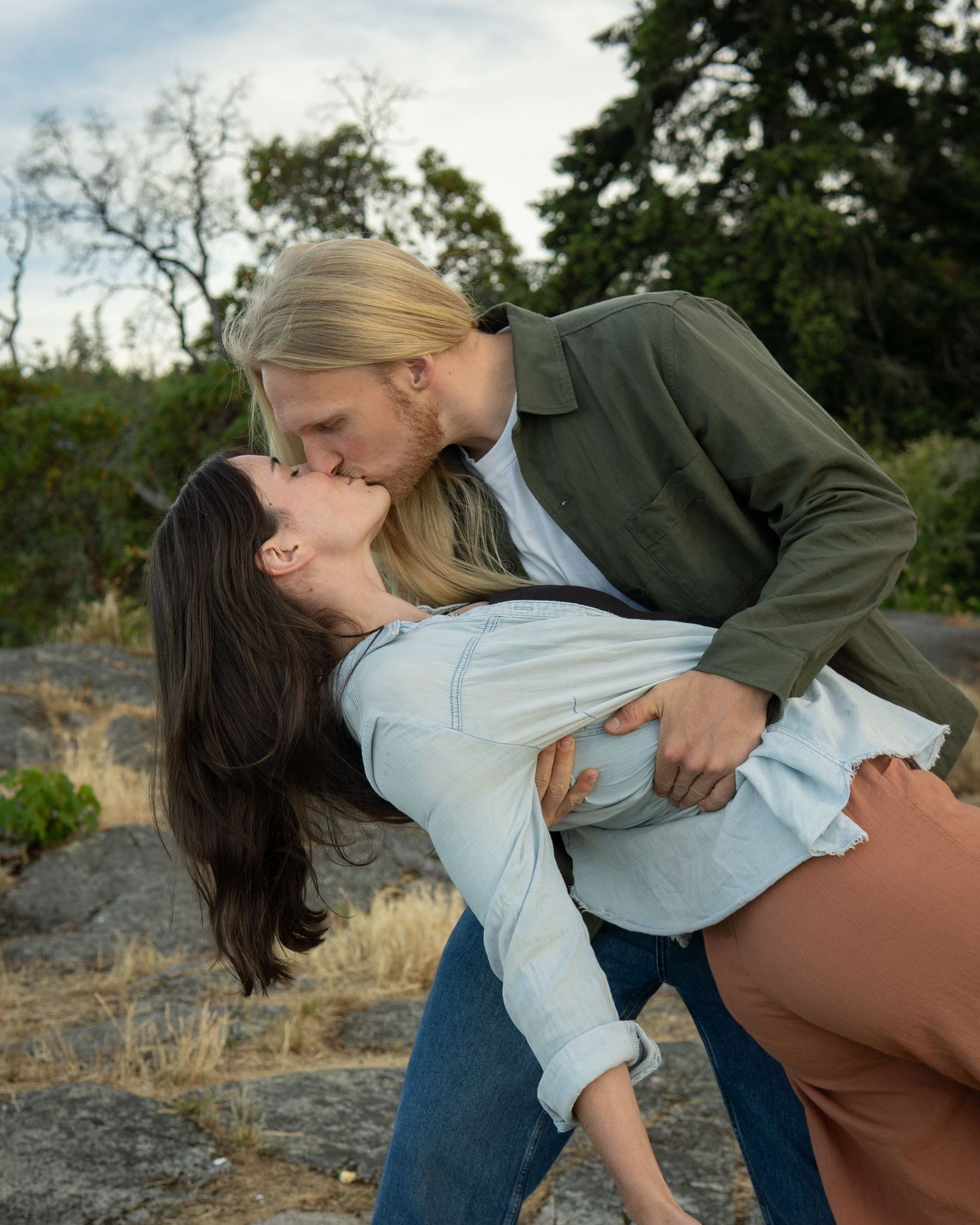 A couple shares a romantic dip kiss outdoors on rocky terrain with trees in the background.