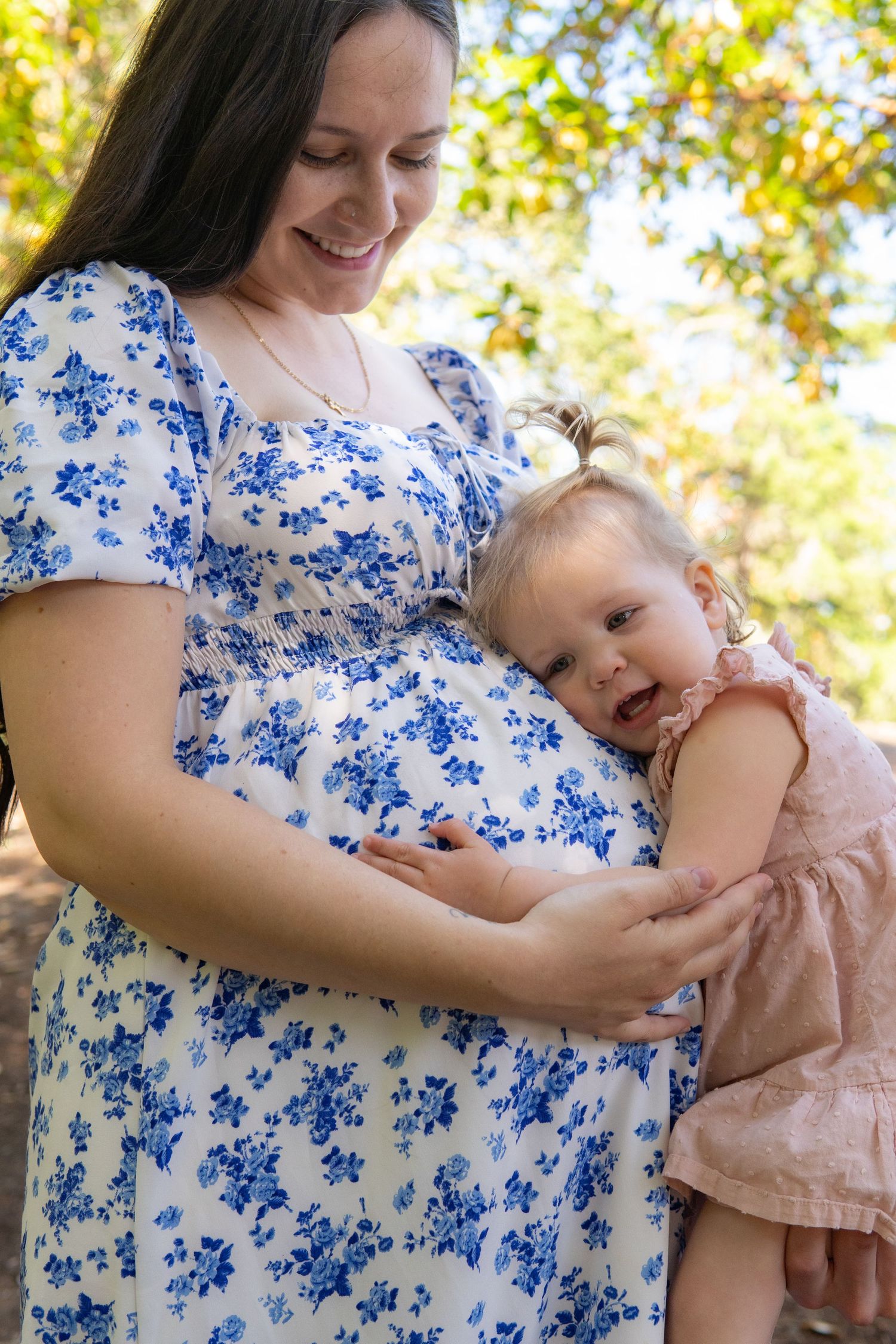 Pregnant woman in floral dress holding toddler girl who rests head on baby bump outdoors.