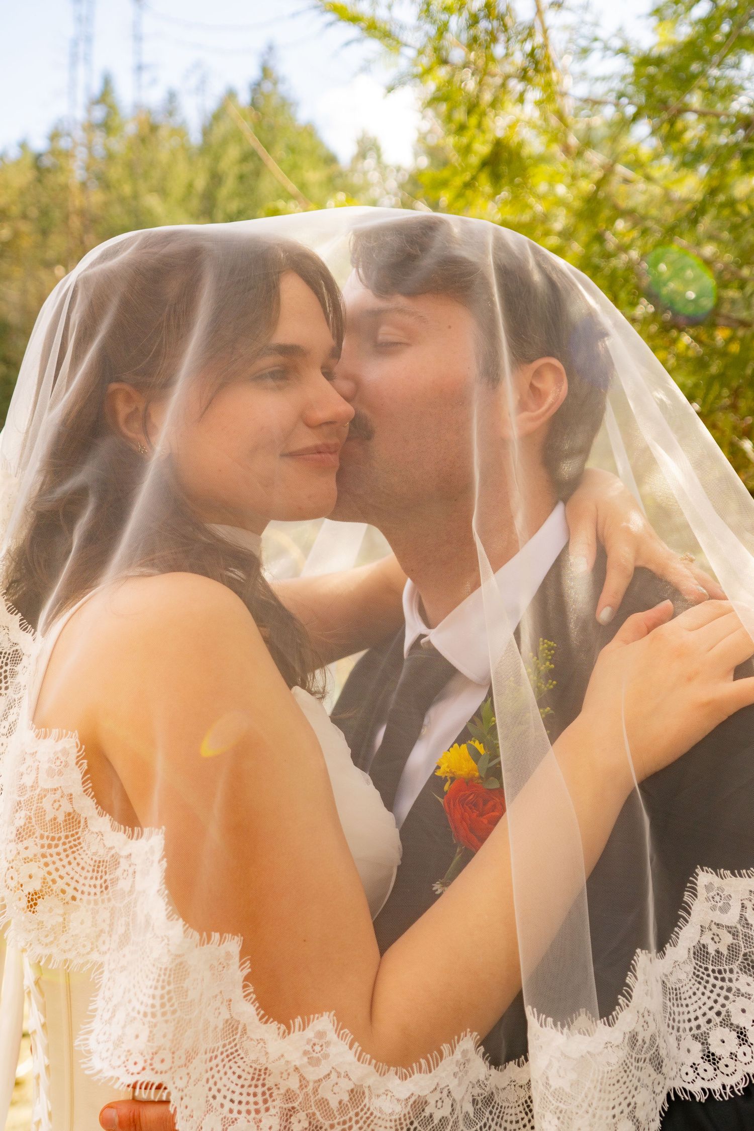 Bride and groom sharing a kiss under a white wedding veil outdoors surrounded by green trees.