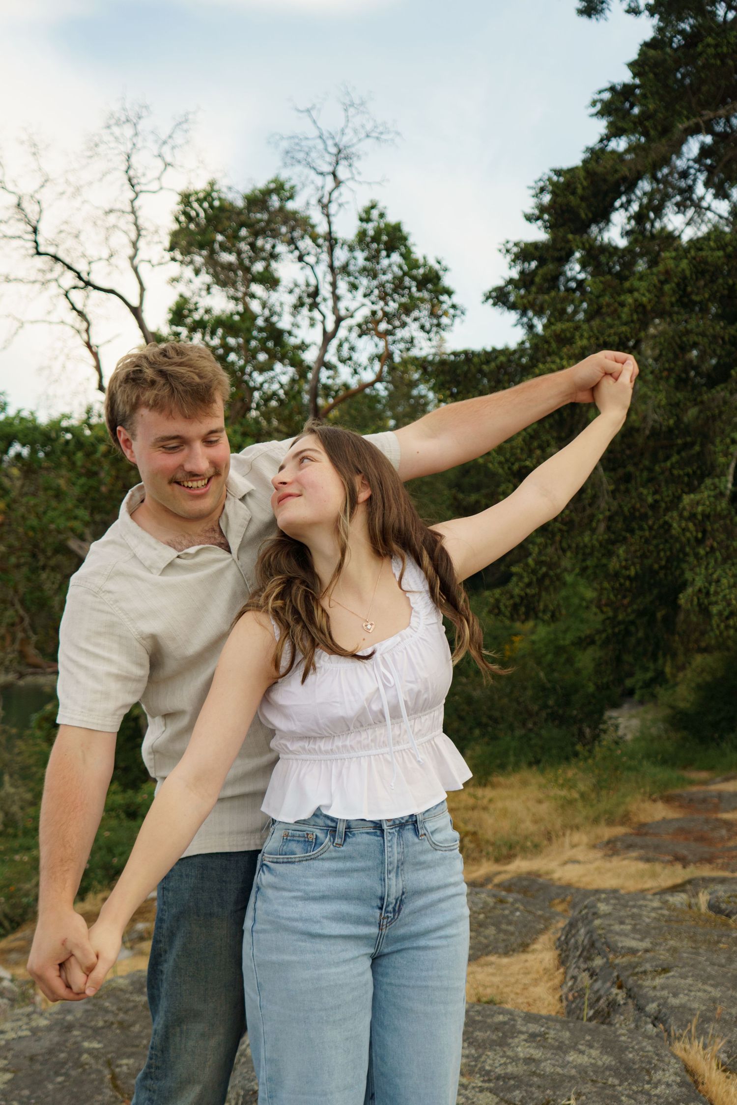 A young couple dancing outdoors, smiling and holding hands, with trees and a cloudy sky in the background.