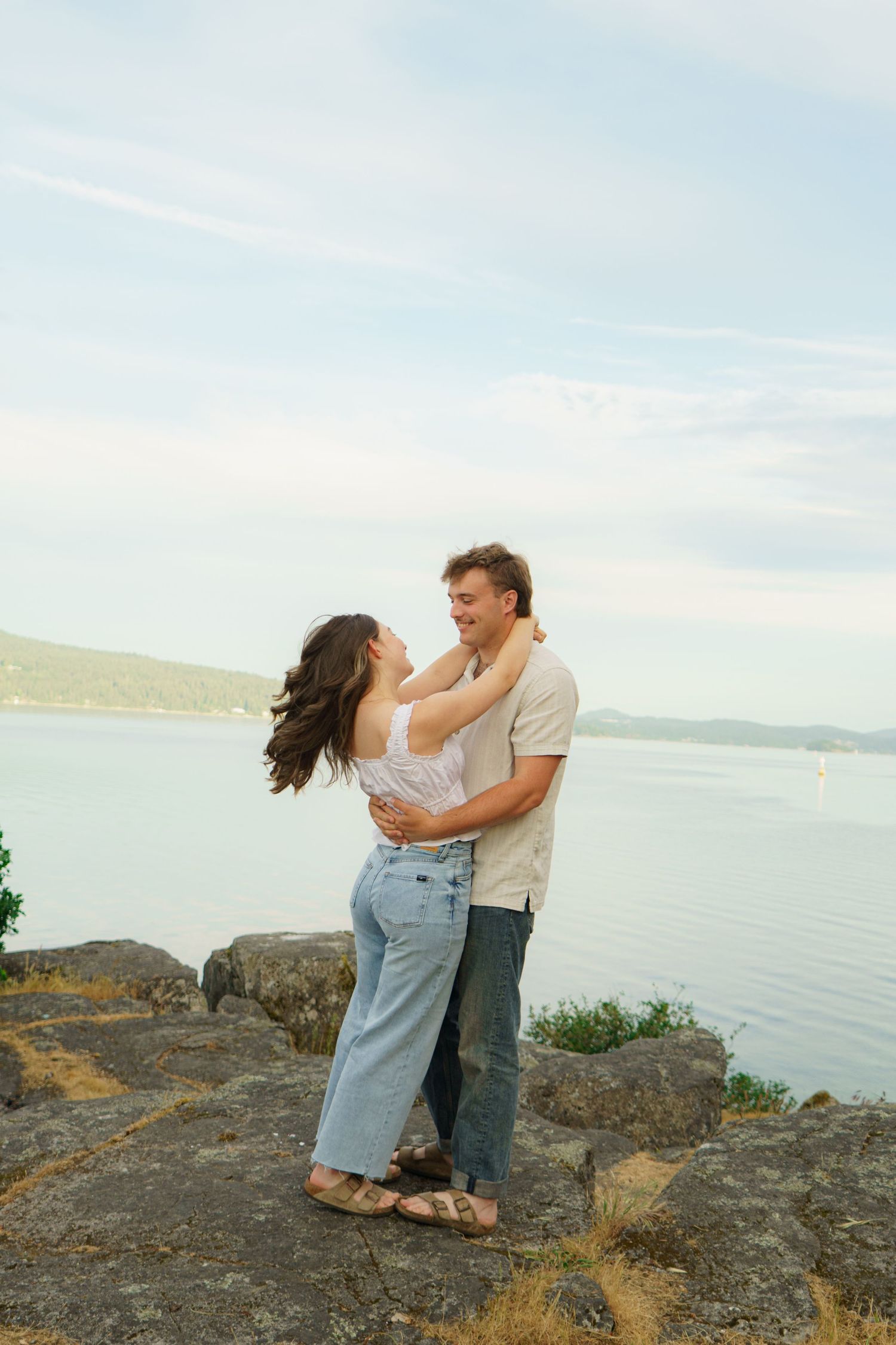 Couple embracing and smiling on rocky shoreline with calm blue water and hills in background.