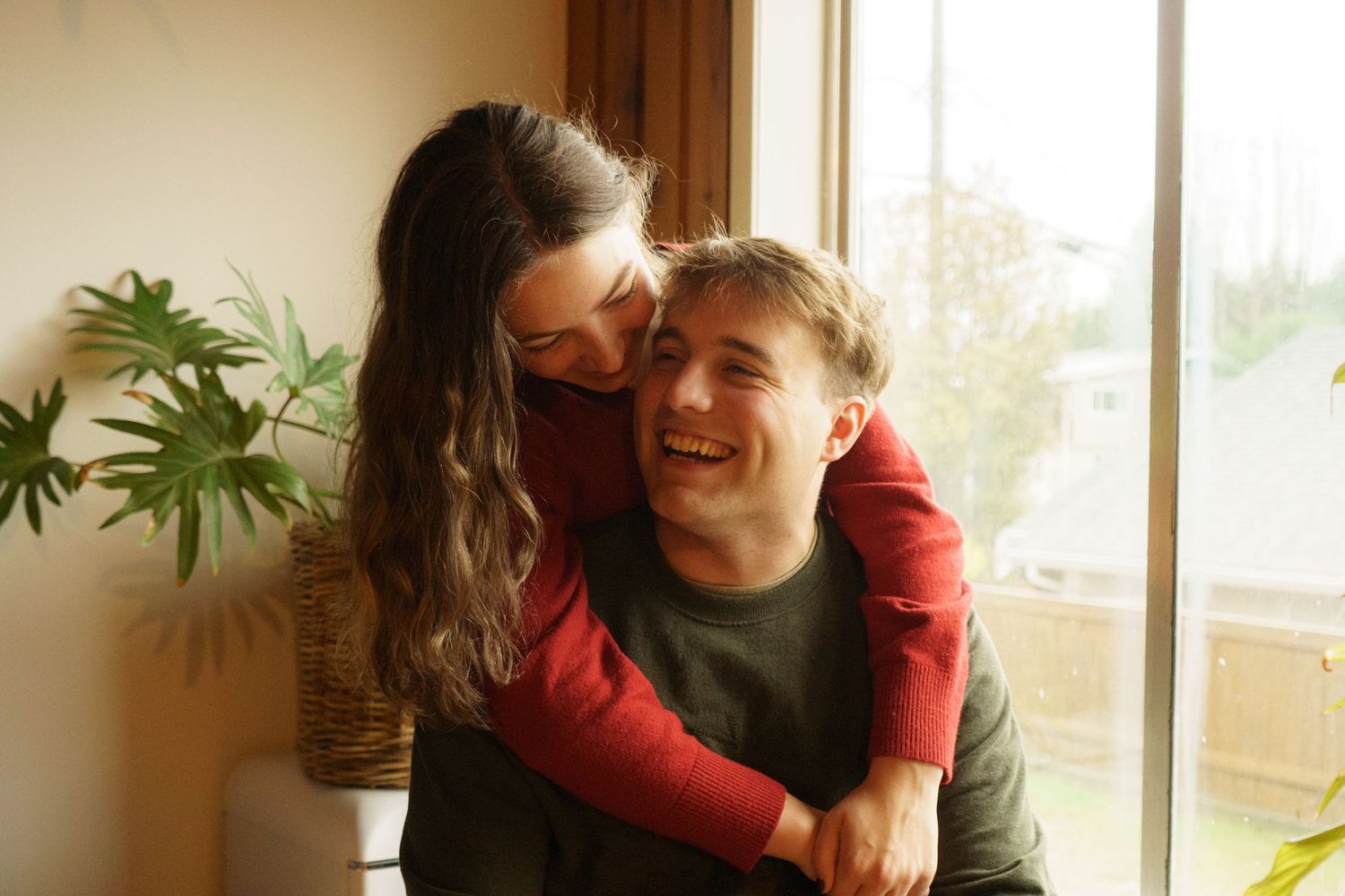 Happy couple laughing and embracing near a bright window with green houseplants in background.