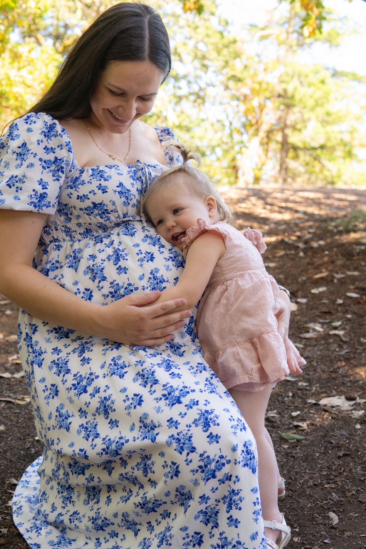 Pregnant woman in floral dress smiling as toddler girl in pink romper hugs her baby bump outdoors.